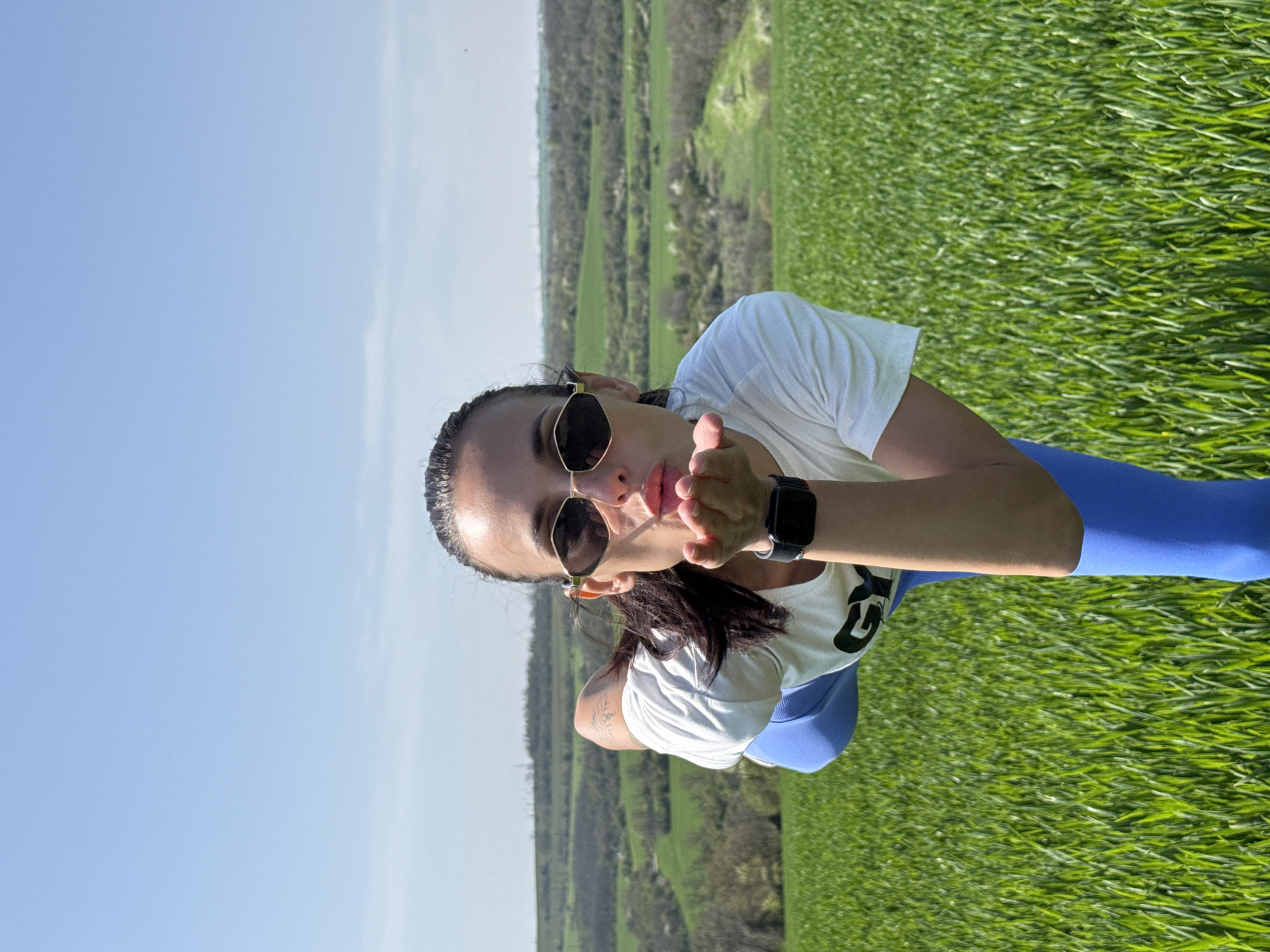 A woman is captured in a vibrant outdoor setting, likely a field in Pleven, Bulgaria, under a bright, clear blue sky. She is positioned centrally, facing the camera with a playful expression, blowing a kiss with her lips puckered. She wears dark sunglasses, a white short-sleeved t-shirt with a partial logo or design (possibly a stylized "G") on the front, and blue athletic leggings. Her dark hair is pulled back, and a dark wristwatch is visible on her left wrist. A tattoo with an abstract, possibly tribal, design is visible on her left upper arm.
The background features a sprawling landscape dominated by lush, bright green grass in the foreground, suggesting a fertile field or meadow. Beyond this, a valley unfolds with more green vegetation and scattered trees, indicating rolling hills or a rural environment. The scene is well-lit by bright daylight, suggesting a clear, sunny day, likely during the late morning or early afternoon. The overall atmosphere conveyed is cheerful and connected with nature.