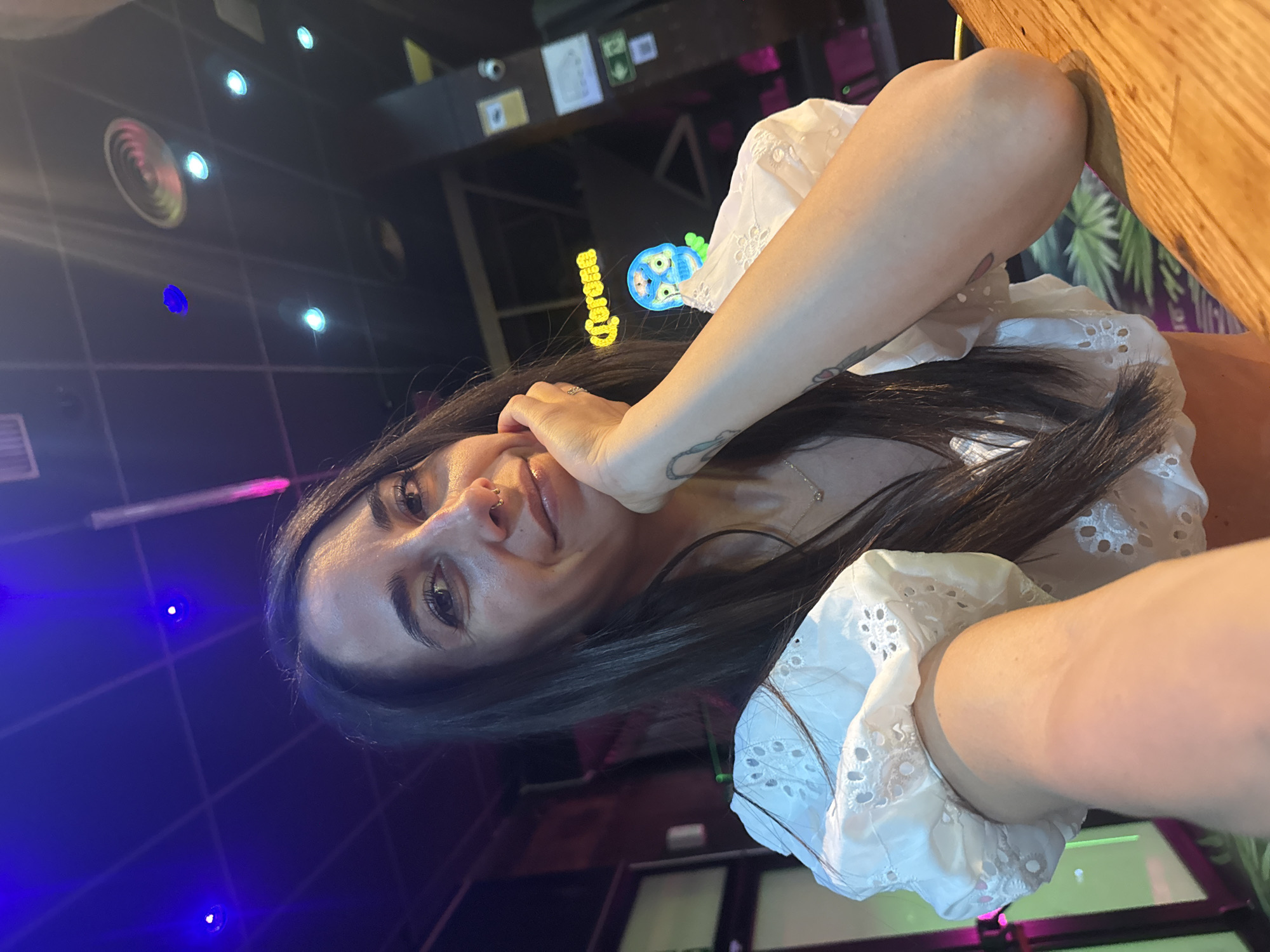 A woman with long dark hair and a nose ring is taking a selfie in what appears to be a bar. She is wearing a white broderie anglaise top. Her right hand is resting on her chin. In the background, a neon "Corona" sign and a blue skull figure are visible. The ceiling has recessed lighting. A wooden table is in the foreground.