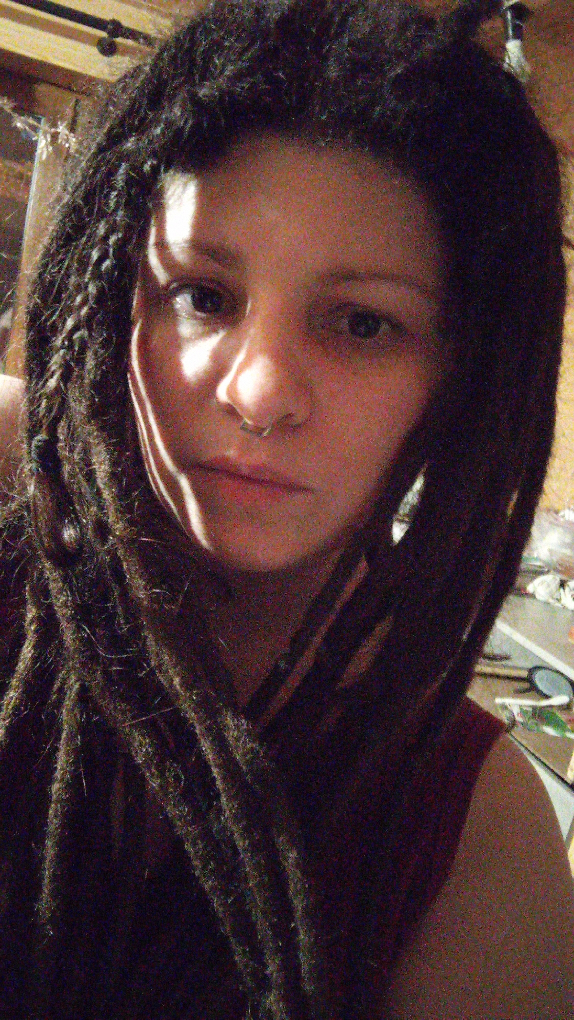 A close-up, slightly grainy photograph depicts a person with dark dreadlocks and a nose ring. Part of the person's face is illuminated by a harsh light. The person appears to be wearing a dark red or maroon shirt. The background is indistinct but suggests an interior space with cluttered surfaces. The presence of the city of Юдино, Russia is not discernible.