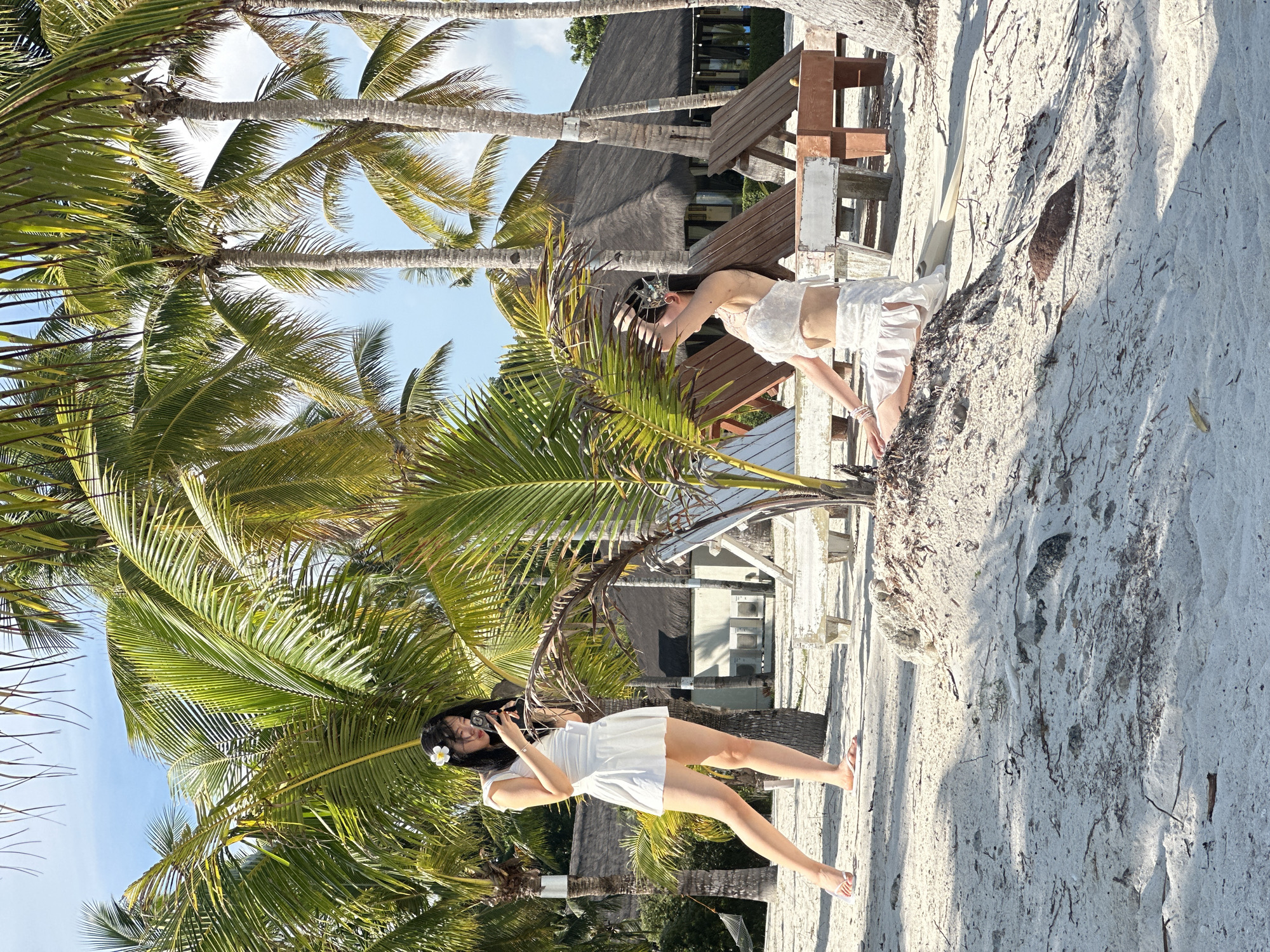 This is a full-shot, eye-level, outdoor photograph of two young Asian women on a sandy beach in Panglao, Philippines. The woman on the left stands facing right, holding a small black camera and looking down at it. She wears a white sleeveless dress and white flip-flops. A white flower is pinned to her dark hair. She is positioned among palm trees. The woman on the right is seated on a mound of sand, facing left with her head tilted back and her right hand raised to her hair. She wears a white two-piece outfit consisting of a cropped top and a ruffled skirt. Behind the women are several palm trees, wooden lounge chairs, and a building with a thatched roof. The ground is covered in white sand and scattered debris. The sky is blue with some clouds.