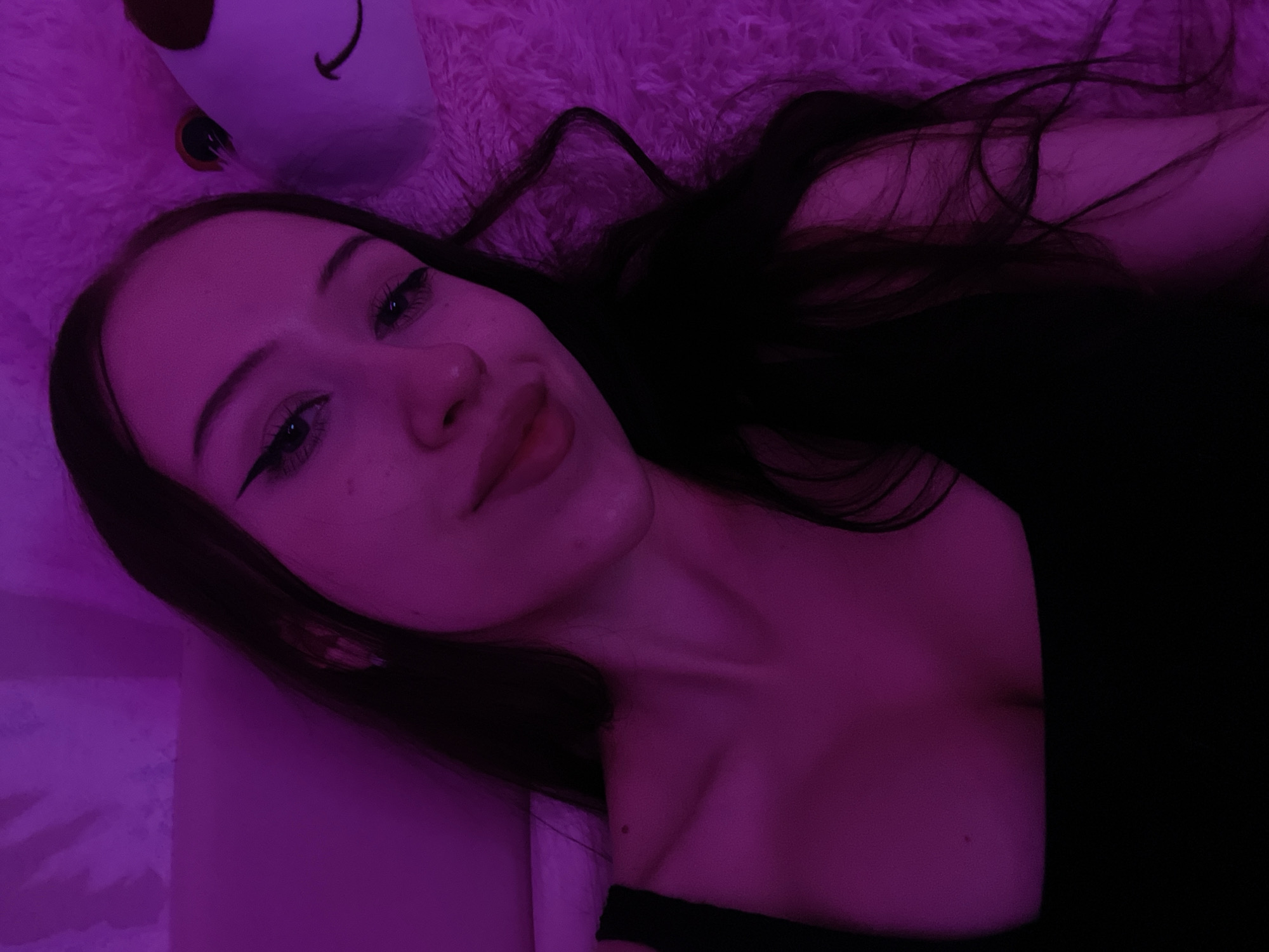 The image is a close-up, low-angle selfie of a young woman with long, dark hair and dramatic winged eyeliner. She is wearing a black, square-neck top. She is illuminated by purple lighting, which casts a strong glow on her skin and surroundings. To her right, a portion of a plush, pink toy is visible, featuring a cartoonish face with round eyes and a smiling mouth. The background appears to be a wall with a textured surface, also under the purple light. The overall impression is of a personal, candid moment captured in a bedroom or similar intimate setting. There is no indication of the city of Votkinsk, Russia.