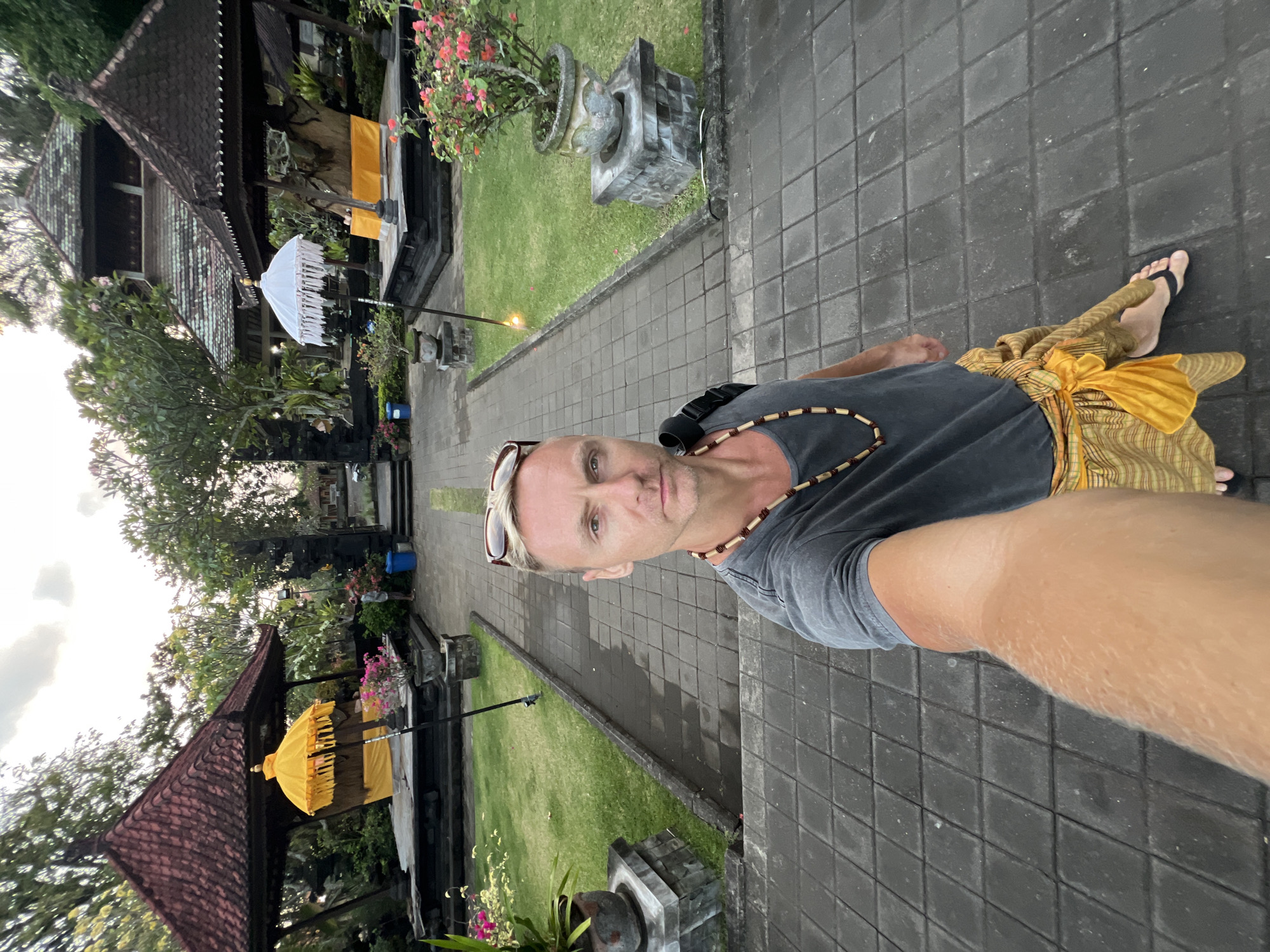In the city of Dawan, Indonesia, a man takes a selfie in front of a temple. The man has blond hair and is wearing a gray t-shirt, a beaded necklace, and a sarong. He is holding a camera with his right arm, which is extended. In the background, there are two traditional Balinese gazebos with ornate roofs. One gazebo is adorned with a bright yellow umbrella and yellow fabric, while the other has a white umbrella and yellow fabric. Both gazebos are situated on raised platforms and surrounded by lush greenery. A stone pathway with square tiles leads through the courtyard, connecting the entrance to the gazebos and the main temple structure visible in the distance. The sky is overcast, suggesting it may be early morning or late afternoon.
