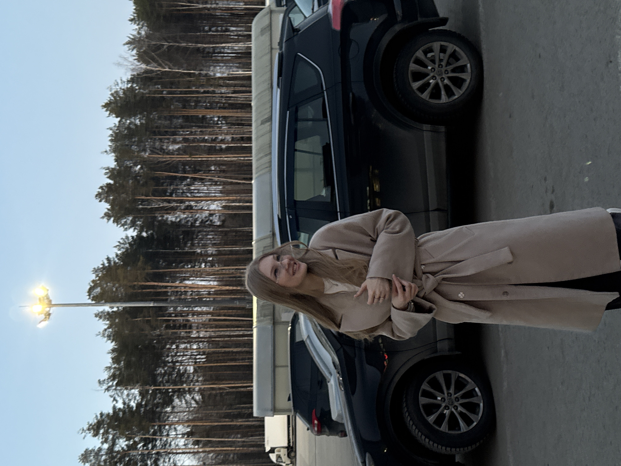 A woman in a beige trench coat stands next to a dark-colored SUV in what appears to be a parking lot. The background features a row of trees and streetlights, suggesting an urban or suburban environment. The scene may be in Perm, Russia. The woman appears to be smiling.