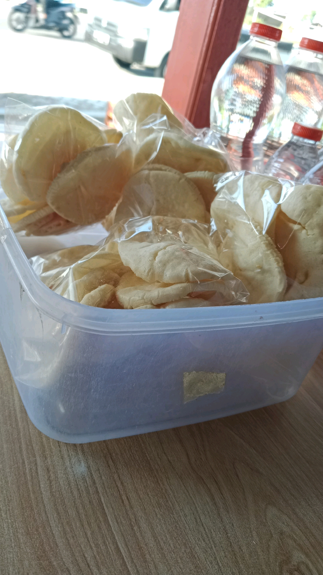 In Sukowinangun, Indonesia, a clear plastic container filled with individually plastic-wrapped rounds of what appear to be crackers or crisps sits on a wooden surface. Behind the container, two plastic bottles of water with red caps are visible. The background is blurred but shows a street scene with a motorcycle in motion and a white vehicle. The lighting suggests it is daytime.