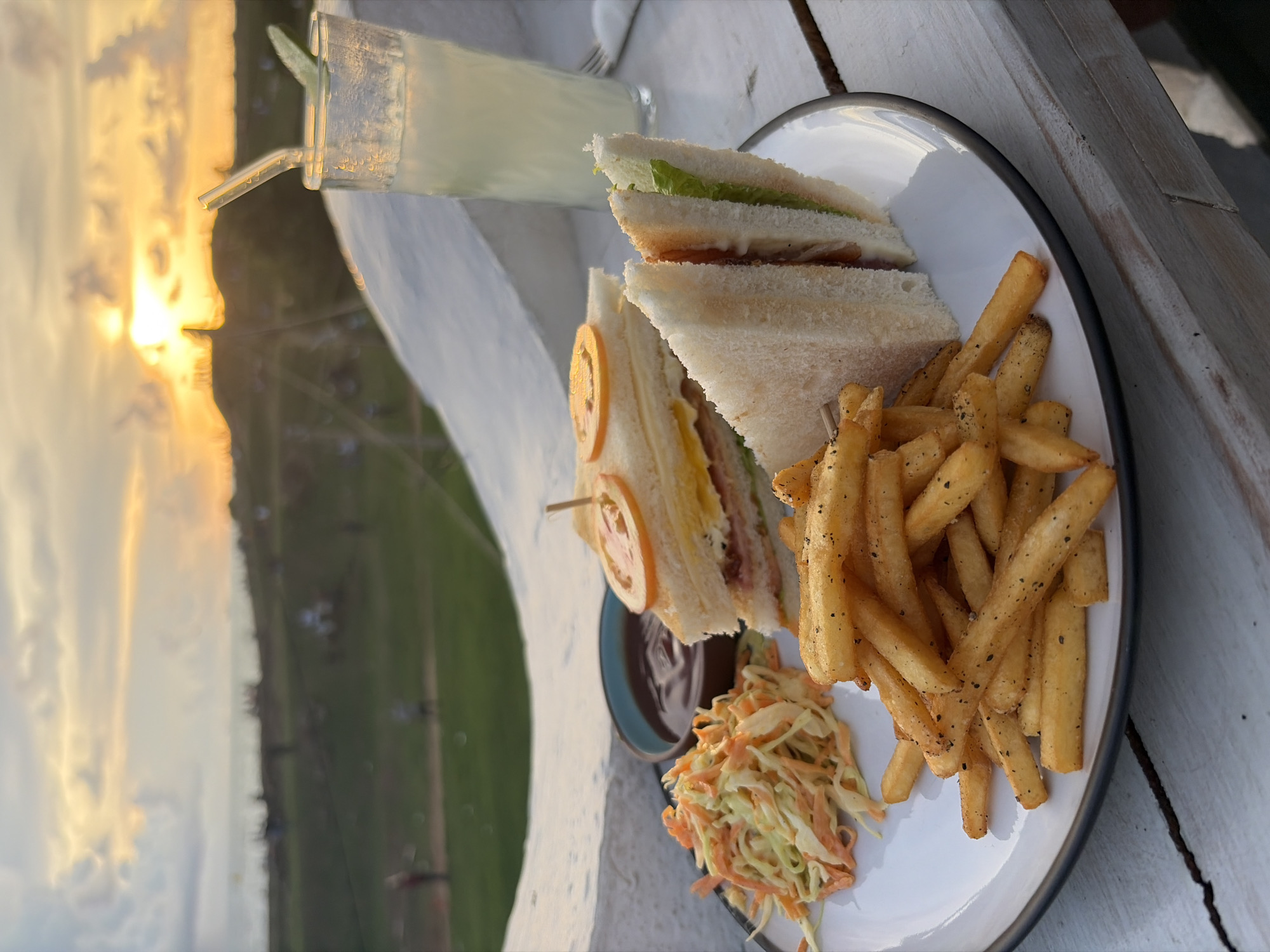 This is an outdoor image of a meal placed on a wooden surface. A white plate with a dark gray trim contains two halves of a sandwich, a pile of french fries, and coleslaw. A small dish with a dark sauce sits nearby. A glass filled with a light-colored liquid, possibly lemonade, with a straw, is also present. The scene overlooks a landscape featuring green fields and a sunset or sunrise. This likely depicts a meal at a location in Galle, Sri Lanka, based on contextual clues. No people are visible in the frame.