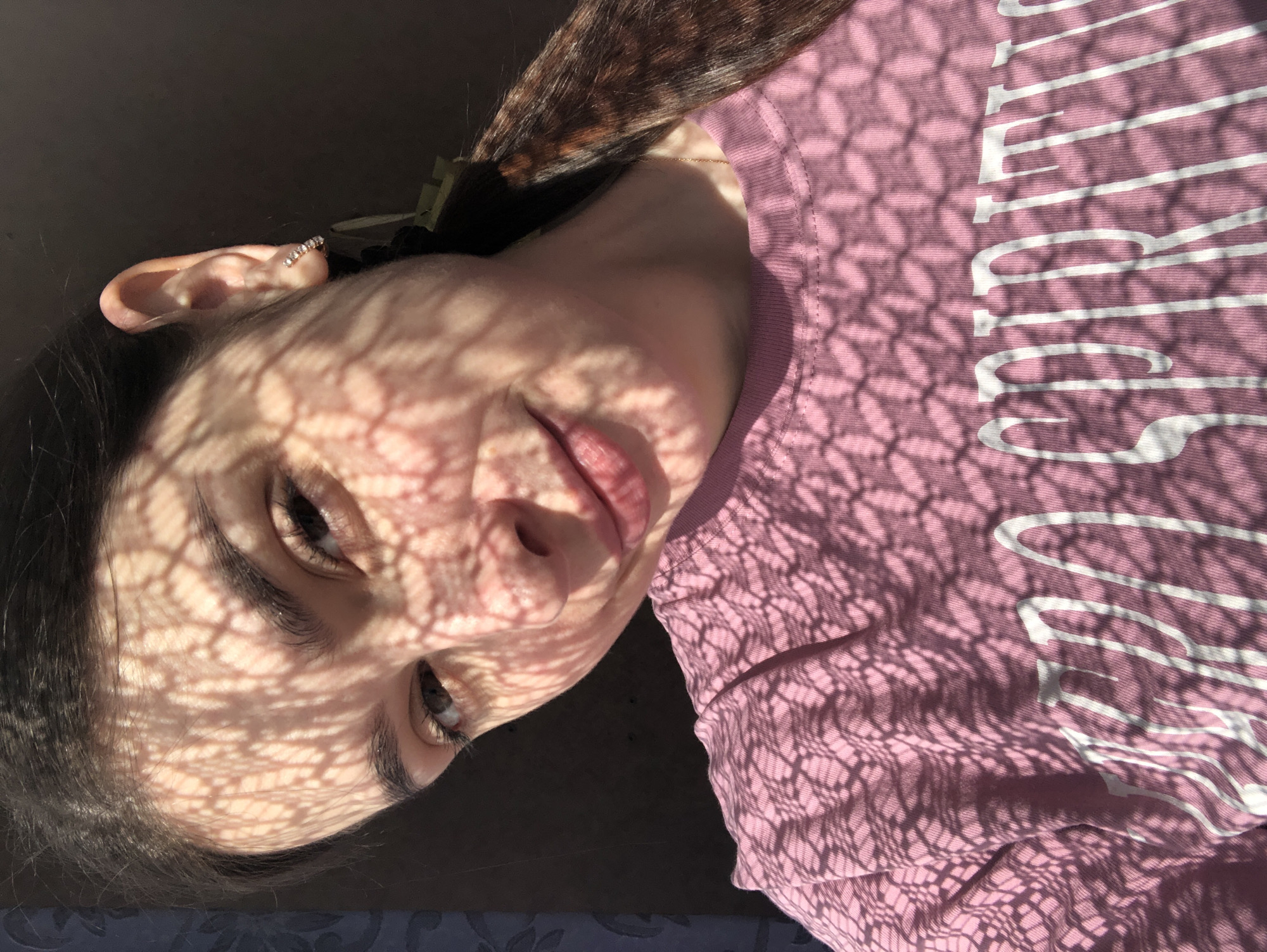The image shows a person with dark hair, light skin, and brown eyes, positioned at an angle. The person has an earring in their left ear. Their face and a portion of their upper body are covered in a pattern of light and shadow, resembling a grid. They are wearing a pink shirt with white lettering. The location is not discernable from the image.
