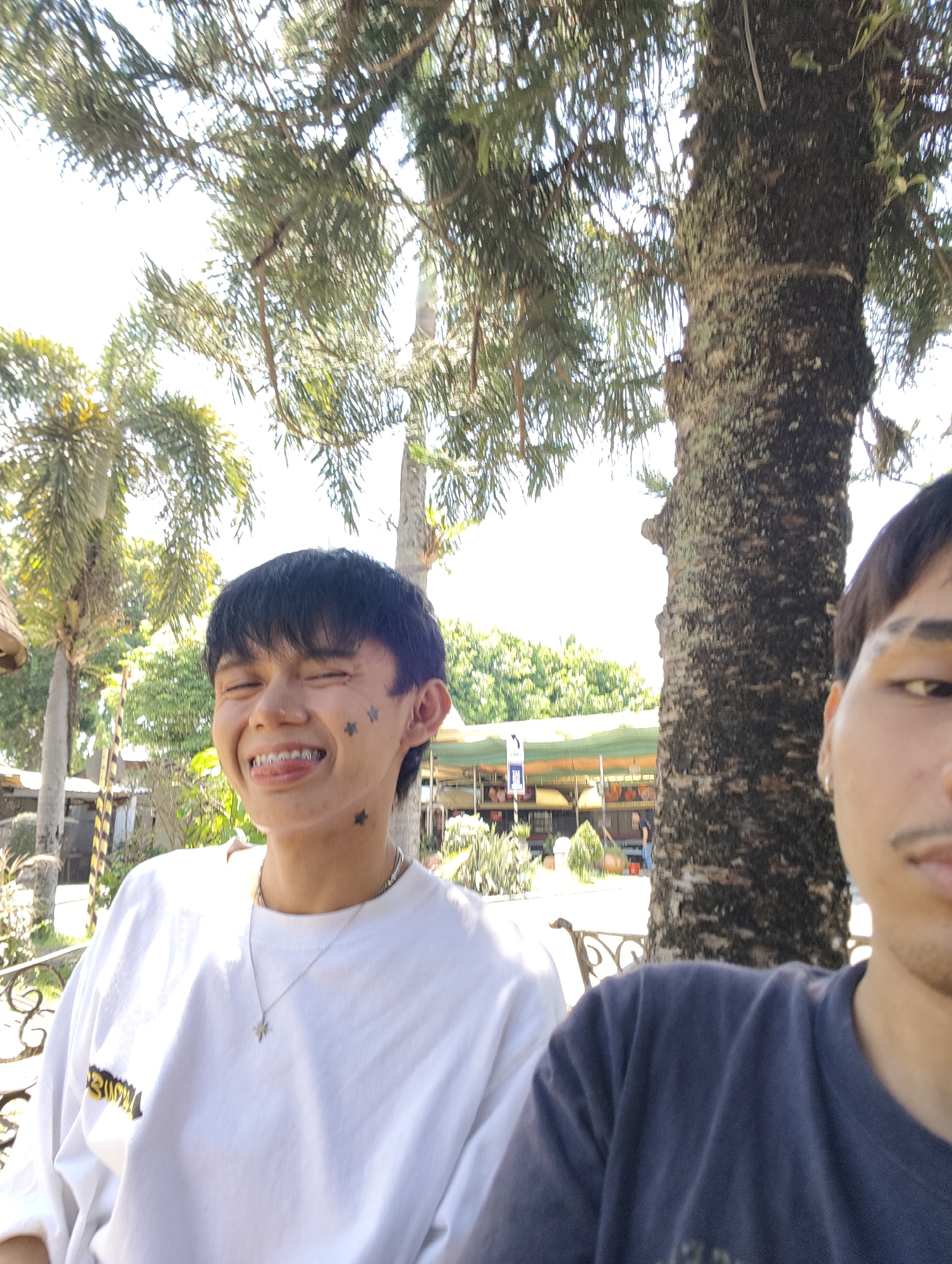 In a sunny outdoor setting in Davao City, Philippines, a young man with short black hair and braces is captured in a selfie. He is wearing a white t-shirt with a star pendant necklace and has star tattoos on his face. He is smiling with his eyes closed and tongue sticking out, showing off his braces. To his right, partially visible, is another person wearing a dark t-shirt. The background features lush green foliage, palm trees, and what appears to be a market or outdoor dining area with awnings and structures. A large tree trunk with textured bark is prominent in the mid-ground, with dappled sunlight illuminating the scene.