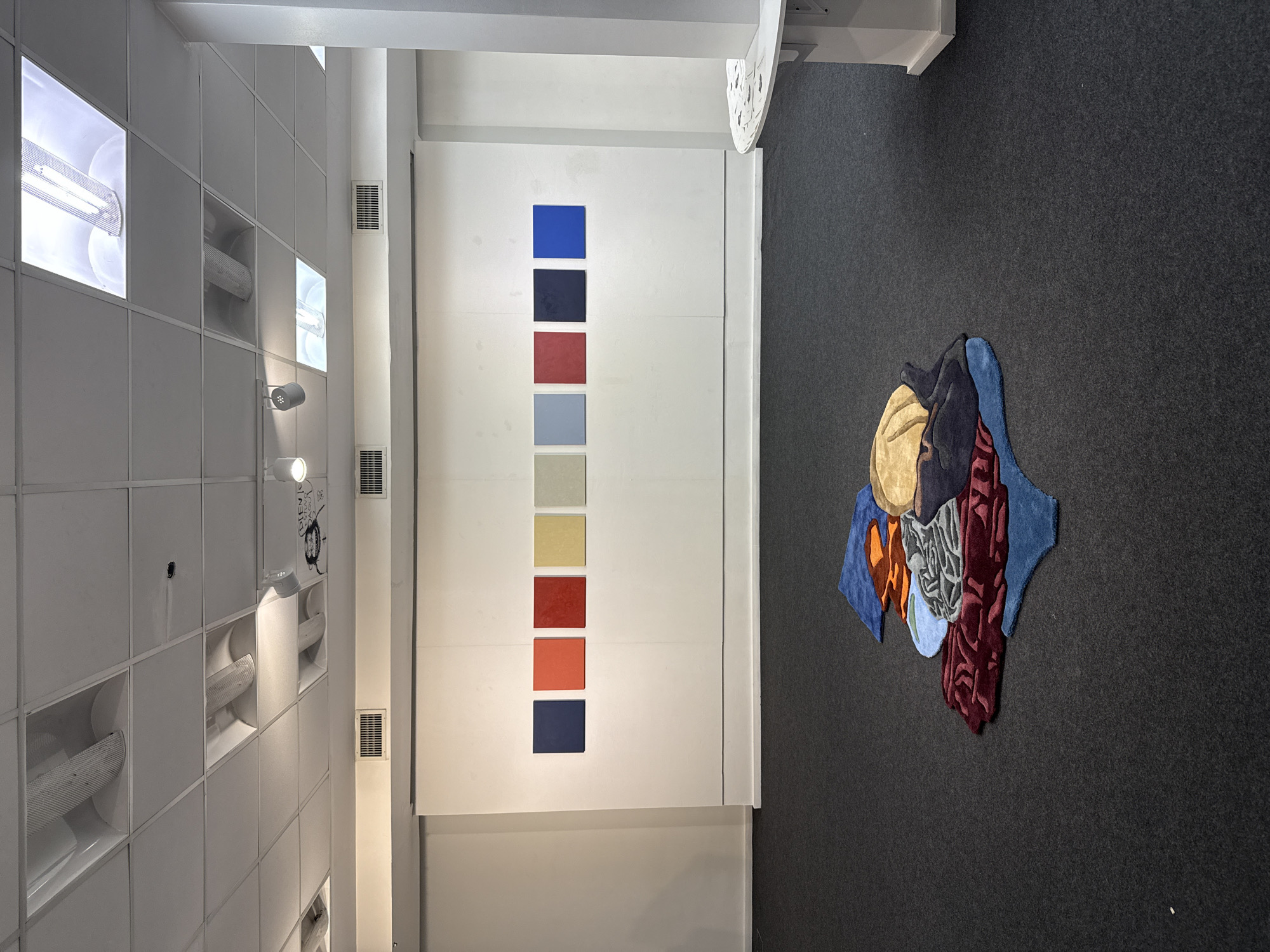 Indoor setting. The scene features a display with a color palette on a white wall, and a rug with an abstract design on a gray floor. The wall on the left has square panels, some containing lights and vents. The color palette consists of nine squares, arranged vertically. From top to bottom, they are blue, dark blue, red, pink, light blue, gray, yellow, orange and dark blue. The rug has shapes of different colors, including blue, gold, red, and gray, giving it an abstract design. The setting could be an art gallery or a design studio. There is no evidence to identify the specific location in Buenos Aires, Argentina.