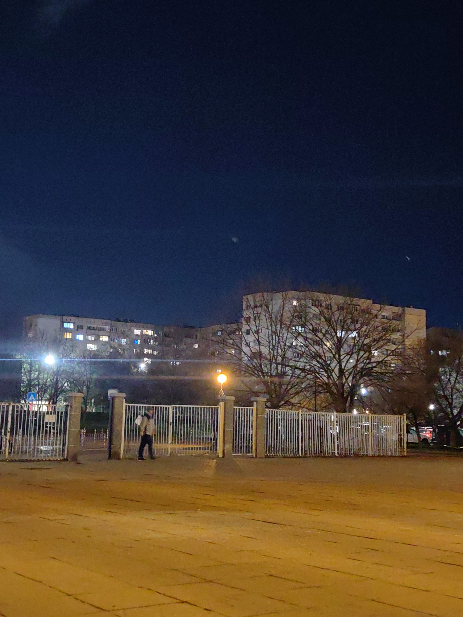 The image displays a nighttime urban scene within a residential area of Sofia, Bulgaria. The foreground features a flat, warm-toned ground surface, likely paved. A white metal picket fence, supported by concrete pillars, runs across the mid-foreground, partially open in the center. Behind this fence, a single individual, wearing a light-colored jacket and dark trousers, walks from left to right, carrying a light-colored object over their left shoulder. The midground includes a public green space with several bare deciduous trees. Multiple artificial light sources are present; a prominent orange streetlamp stands on the right, and a brighter, whiter light source is visible on the left behind the fence. A blue and white pedestrian crossing sign is also discernible on the left side, behind the fence. The background is dominated by several large, light-colored, multi-story apartment buildings, characteristic of post-WWII Eastern European architecture, with numerous windows illuminated by internal lights. The sky above is dark blue, with faint atmospheric haze or distant light sources visible.