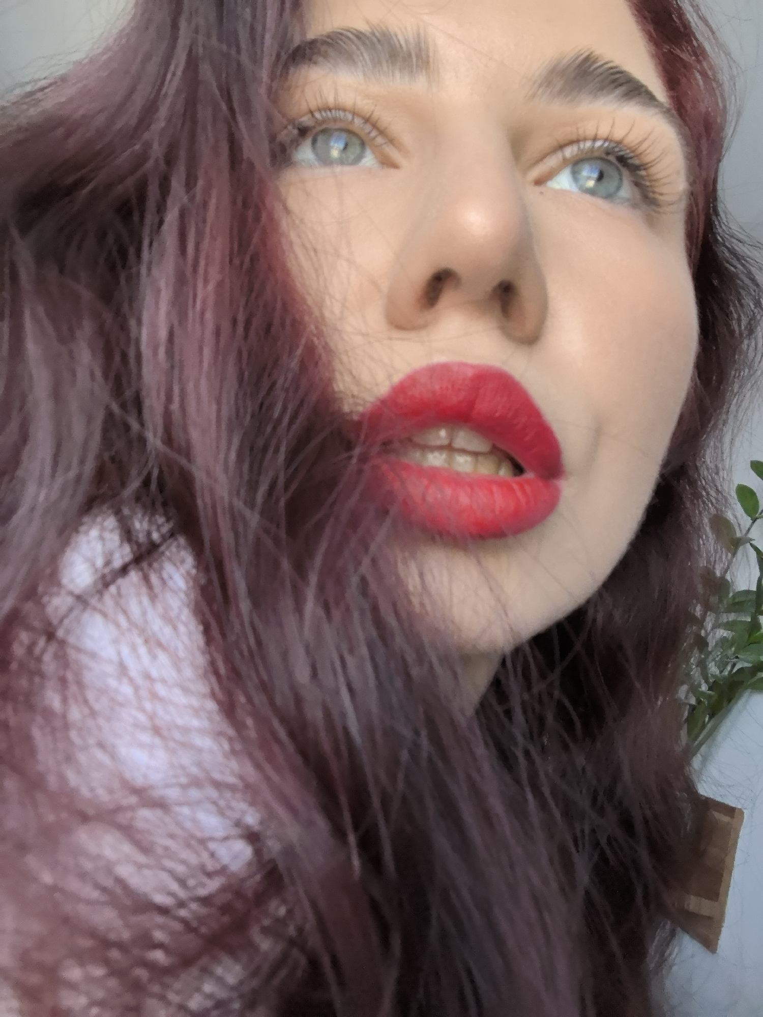 The image is a close-up shot of a woman's face. She has fair skin, light blue eyes, and auburn or burgundy-colored hair that partially obscures her face. Her lips are painted with bright red lipstick and are slightly parted. The woman is looking upwards. The background is blurry, but some greenery is visible on the right side of the frame. There is no information about location (Stara Zagora, Bulgaria) available in the image.