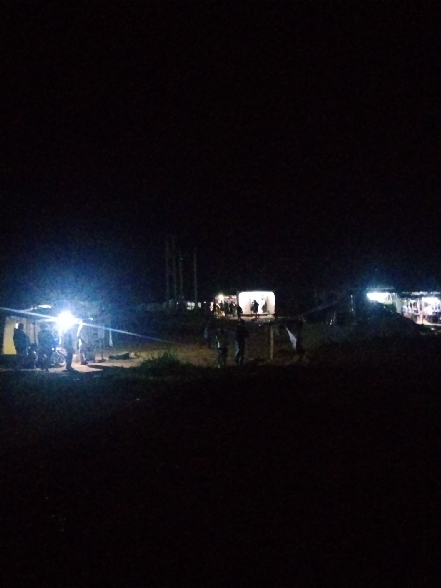 The image depicts an outdoor scene at night in the city of Durumi, Nigeria. There are several people visible, some gathered around what appear to be motorcycles under artificial lighting. In the background, there are structures that might be buildings or stalls, illuminated by lights. The environment is dark, with the primary light sources being artificial, casting bright beams and illuminating the immediate vicinity of the gatherings. The ground appears to be uneven and covered with some vegetation. The overall impression is of a temporary gathering or event taking place after dark in a semi-urban or rural setting within Durumi.