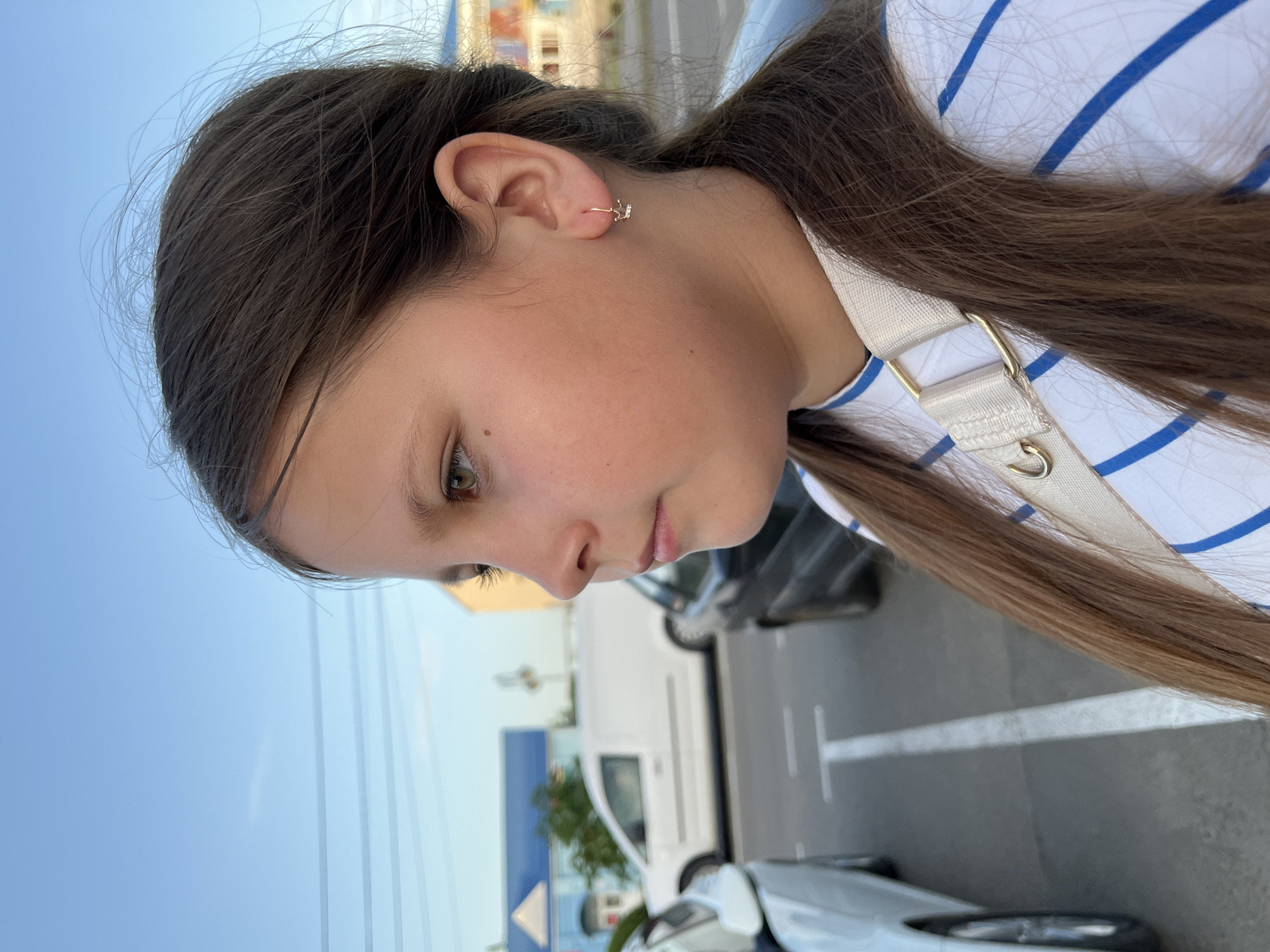 A young girl with brown hair pulled back into a ponytail is seen from a low-angle perspective, gazing downwards. She is wearing a white and blue striped shirt with a white strap across her shoulder. A gold earring is visible in her ear. The background suggests an outdoor parking area with vehicles, including a white van. A building, possibly a store, with blue and white signage is also visible. Based on the available information, it is not possible to confirm the exact location as Novopokrovskaya, Russia.