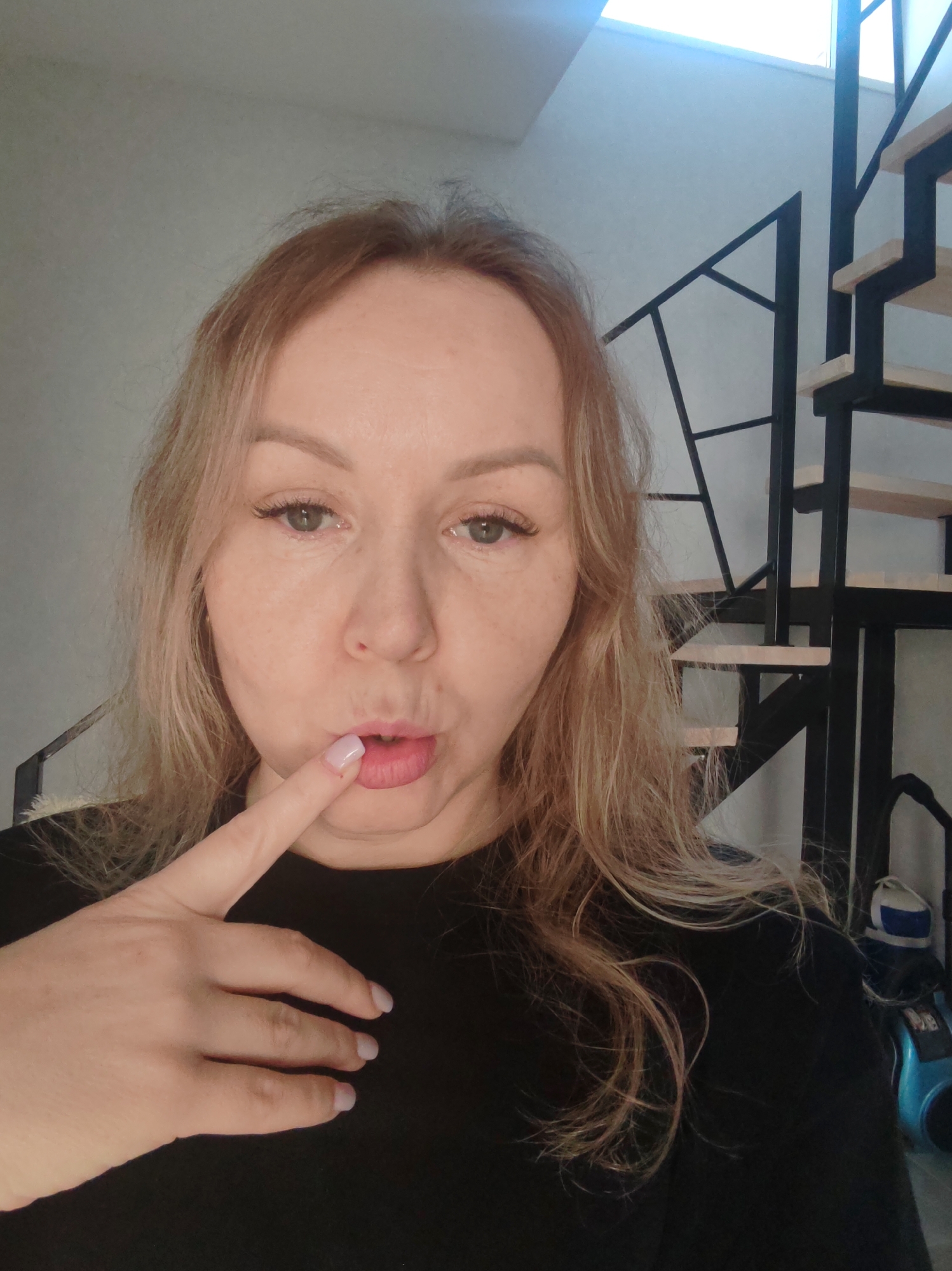 A woman with fair skin and blonde hair poses for a selfie indoors. She is wearing a black shirt and holding her index finger to her lips. Behind her, there is a modern staircase with black metal supports and wooden steps. In the background, a blue and black canister vacuum cleaner is visible. The location is likely in Chaikovsky, Russia.