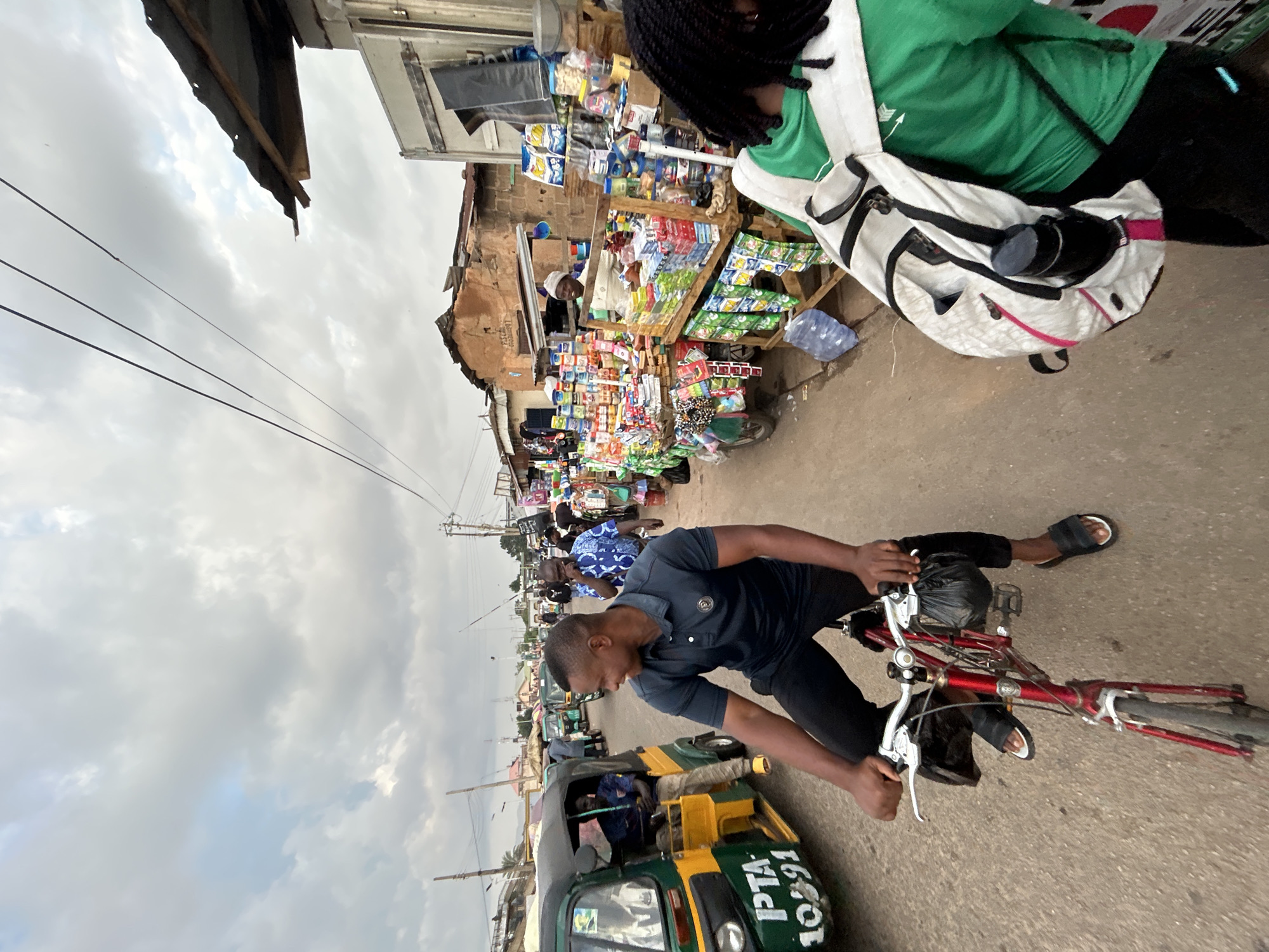 The scene captures a bustling street in Jos, Nigeria. A man in a dark blue shirt and black pants rides a red bicycle along the right side of the road. He wears sandals on his feet. Beside him, there is a green and yellow "Keke Napep," a type of auto-rickshaw, bearing the license plate "PTA-10391" visible. Further ahead, a street vendor has set up a stall overflowing with a variety of goods, mostly snacks and confectionery. A person wearing a green shirt and a white backpack stands near the vendor. In the background, more people and vehicles can be seen lining the street, suggesting a crowded marketplace or shopping area. Overhead, power lines stretch across a cloudy sky. Buildings on either side of the street appear to be a mix of commercial and residential structures, reflecting the urban landscape of Jos.