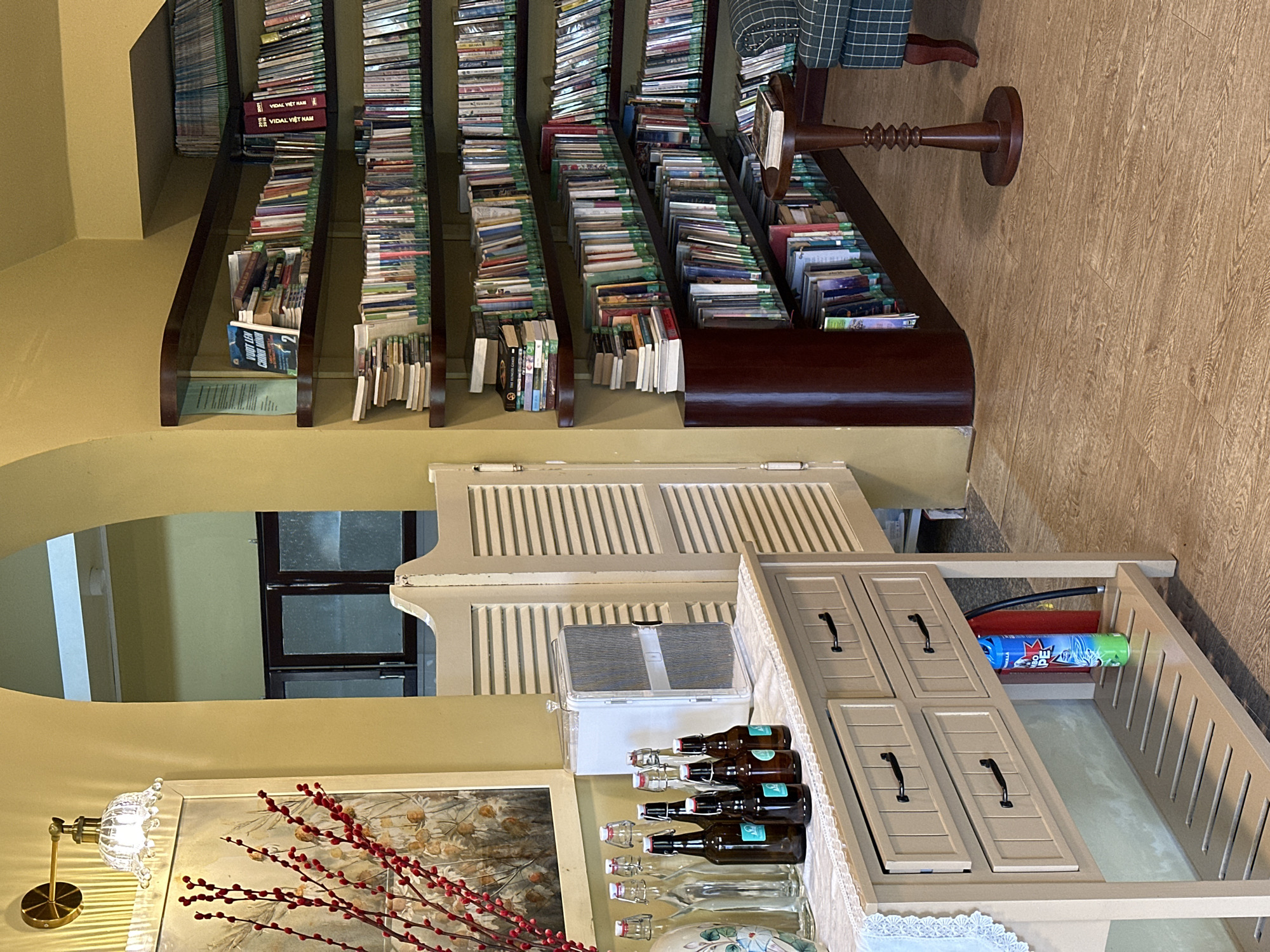 The image depicts an indoor scene, likely a library or study, located in Da Lat, Vietnam. The room is characterized by warm, yellow-toned walls and a wooden floor. Prominent features include extensive bookshelves filled with a variety of books, arranged on dark wood shelves that form a corner. To the left, a white cabinet with drawers stands on a white lace runner, adorned with several glass bottles and brown bottles, some with stoppers. Above the cabinet, a framed artwork featuring red branches is visible, lit by a brass wall sconce. In the background, a white louvered door or divider is present. A small, dark wooden side table with a book on top sits near a green and white checkered armchair in the corner. The overall ambiance suggests a cozy and studious environment.