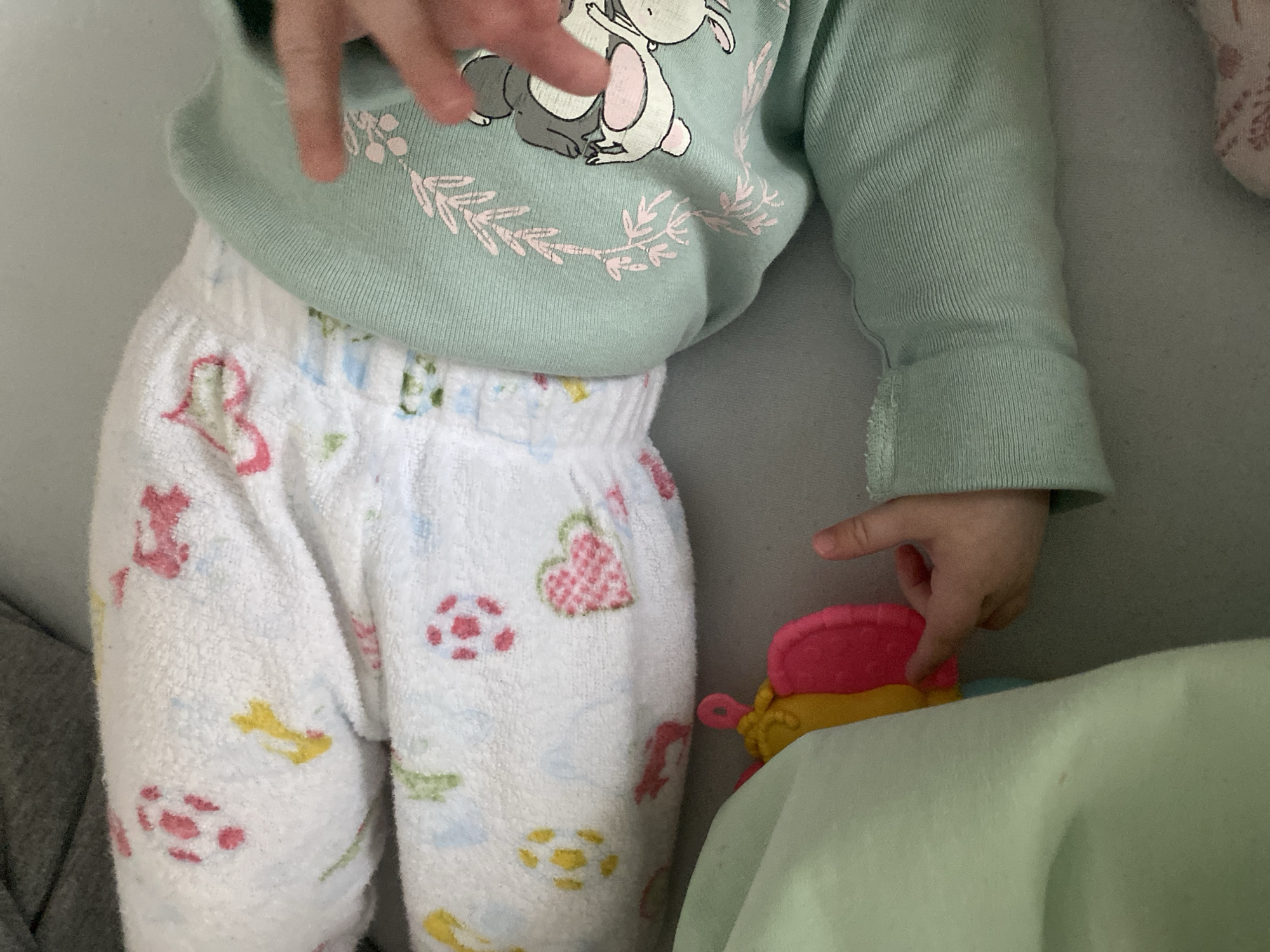A child wearing a light green long-sleeved shirt with a rabbit design and white patterned pajama pants is reaching towards a pink and yellow toy. The location is not discernible from the image. The image depicts a child's clothing and a toy, and the child is interacting with the toy. The environment appears to be indoors, possibly a bed or play area.