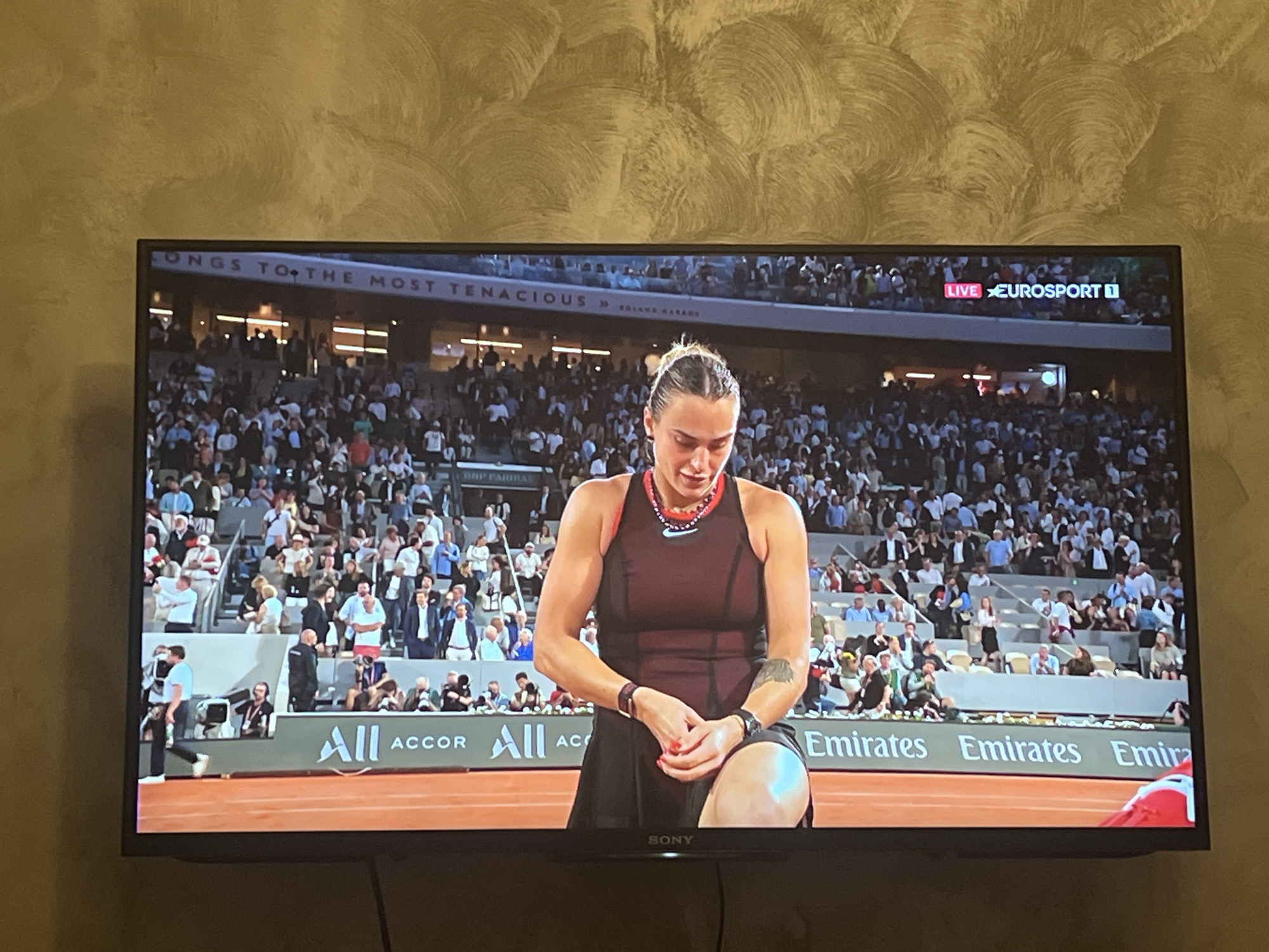 The image displays a television screen broadcasting a live tennis match, situated in a room in Sofia, Bulgaria, with a textured, swirl-patterned wall visible above and beside the TV. The main subject on screen is a female tennis player, wearing a dark sleeveless dress with a red-accented neckline and a dark watch, kneeling on a red clay court. Her head is bowed, and her hands are clasped, suggesting a moment of reflection or intense focus during a break in play.
The background reveals a large stadium filled with spectators, indicating a major professional event. A banner above the stands partially reads "ONGS TO THE MOST TENACIOUS" and "ROLAND GARROS," identifying the venue as the French Open. Advertising boards display "All ACCOR" and repeated "Emirates" logos along the court's edge.
In the top right corner of the TV screen, "LIVE EUROSPORT 1" is visible, confirming the broadcast's live status from the sports channel. The television set itself is a "SONY" brand, as indicated by the logo on its bottom bezel. The match appears to be taking place in the evening or at night under stadium lighting.
