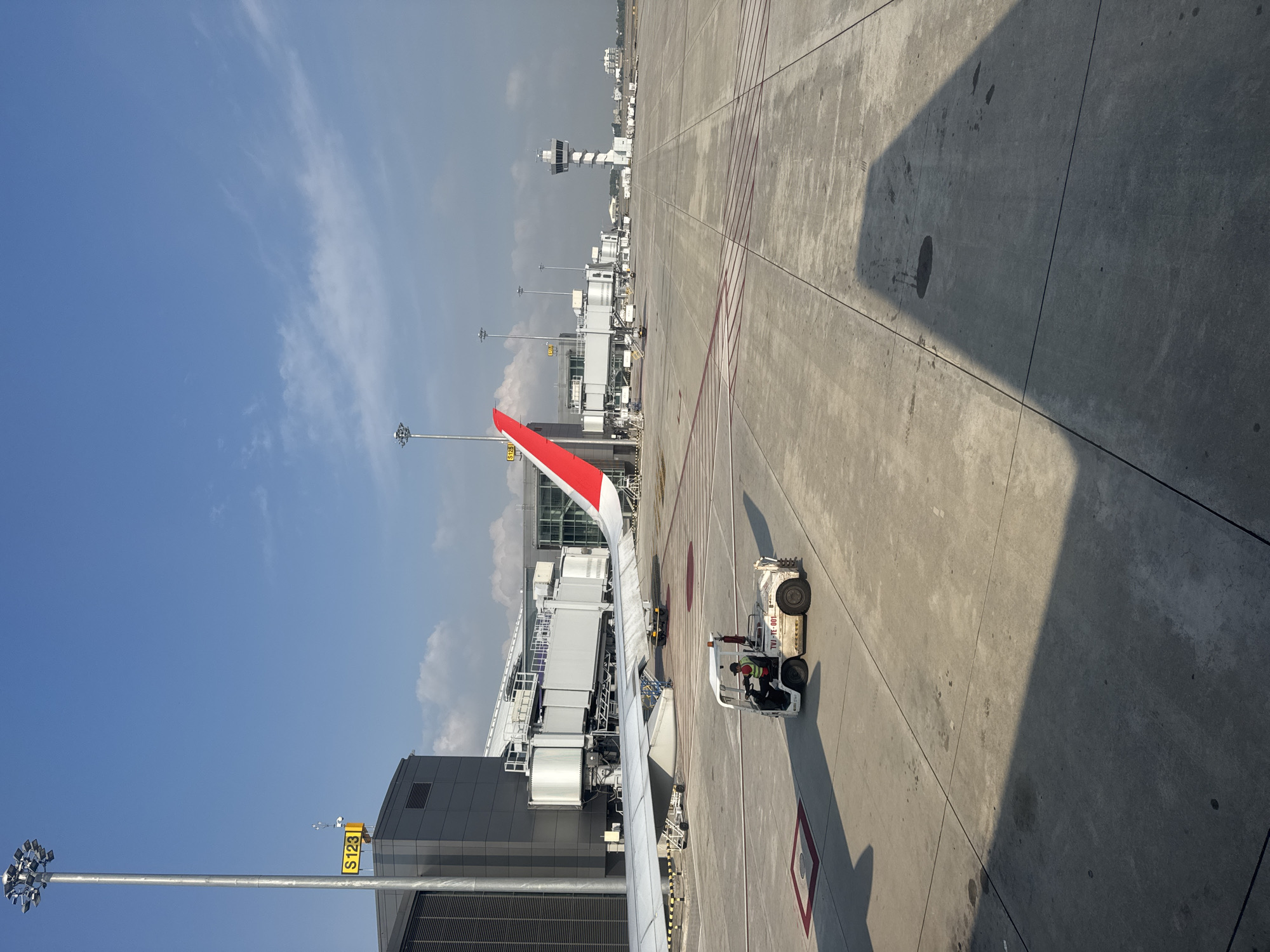 The image depicts an airport apron with an airplane wing visible, a baggage tractor near a jet bridge, and an air traffic control tower in the background. The location is inferred to be in Nong Prue Subdistrict, Thailand. The sky is blue with scattered clouds. There is a sign "S123" on a light pole. There is a shadow cast on the pavement.