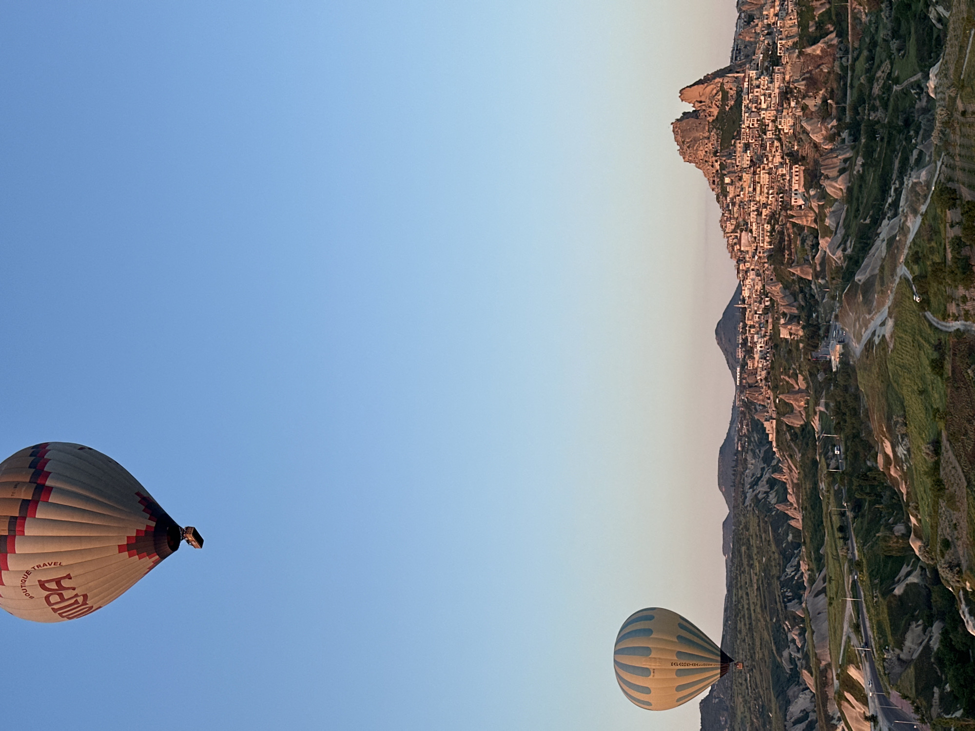 The image, oriented vertically, captures a scenic hot air balloon flight over a unique landscape. The left side of the frame is dominated by a clear sky, transitioning from a vibrant blue at the top to a lighter, warmer blue-yellow hue towards the right, suggesting either sunrise or sunset.
Two hot air balloons are visible. The larger balloon, positioned towards the lower-left, is predominantly white with striking red and black striped patterns. Prominently displayed text on its surface reads "BUTIQUE TRAVEL" and a larger, bold word, likely "TUPA" or "KUPA", in red. A smaller, second hot air balloon appears further to the right and lower in the frame. This balloon is light-colored with distinct blue stripes, and faint letters and numbers, possibly "TC-BOZ", can be seen on its side.
The right side of the image reveals a distinctive landscape. In the mid-ground, a magnificent, multi-peaked rock formation, characteristic of a "fairy chimney," is densely covered with buildings and structures, indicating a unique town or settlement carved into and built upon the rock. This settlement is highly characteristic of Uçhisar, Turkey, aligning with the provided location context. The surrounding terrain consists of rugged hills and valleys with patches of green vegetation, and a winding road or path is visible through the landscape. No people are distinctly visible. The overall scene portrays the serene and majestic activity of hot air ballooning over the historic and dramatic geological formations of Cappadocia during the golden hours of the day.