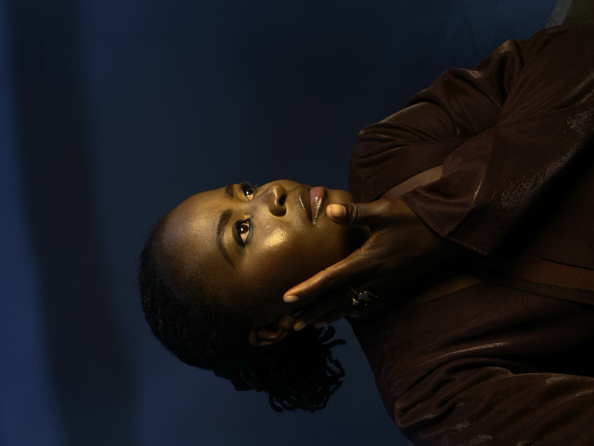 The media file shows a close-up, low-angle shot of a Black woman's face and upper body. She is wearing a shimmering brown jacket. Her head is tilted back and to the left, with her gaze directed upwards. Her left hand is brought up to her face, with her fingers gently touching her chin and cheek. A ring with a dark stone is visible on her left ring finger. The background is a dark, textured blue. The lighting is dramatic, with a warm light highlighting the woman's skin and creating shadows. The overall mood is contemplative and introspective. The image is oriented vertically, as if the woman is lying down or looking up from a lower position. There is no visible text in the image.