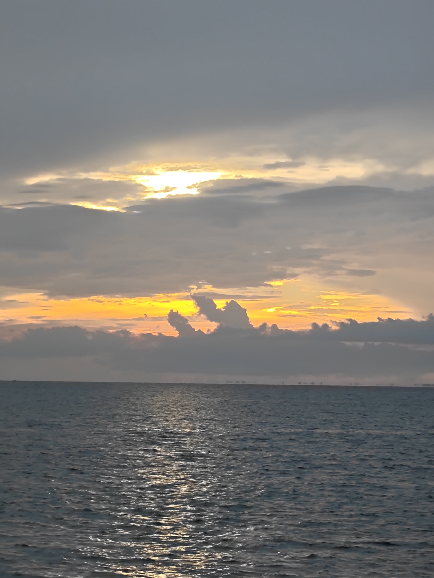 The image depicts a seascape at dusk in Lhokseumawe, Indonesia. The sea is choppy, reflecting the fading sunlight. A horizon line is visible in the distance, with faint suggestions of land or structures. Above, the sky is heavily clouded, with a gap near the horizon where the setting sun casts a bright yellow-orange glow. The light illuminates the edges of the clouds.