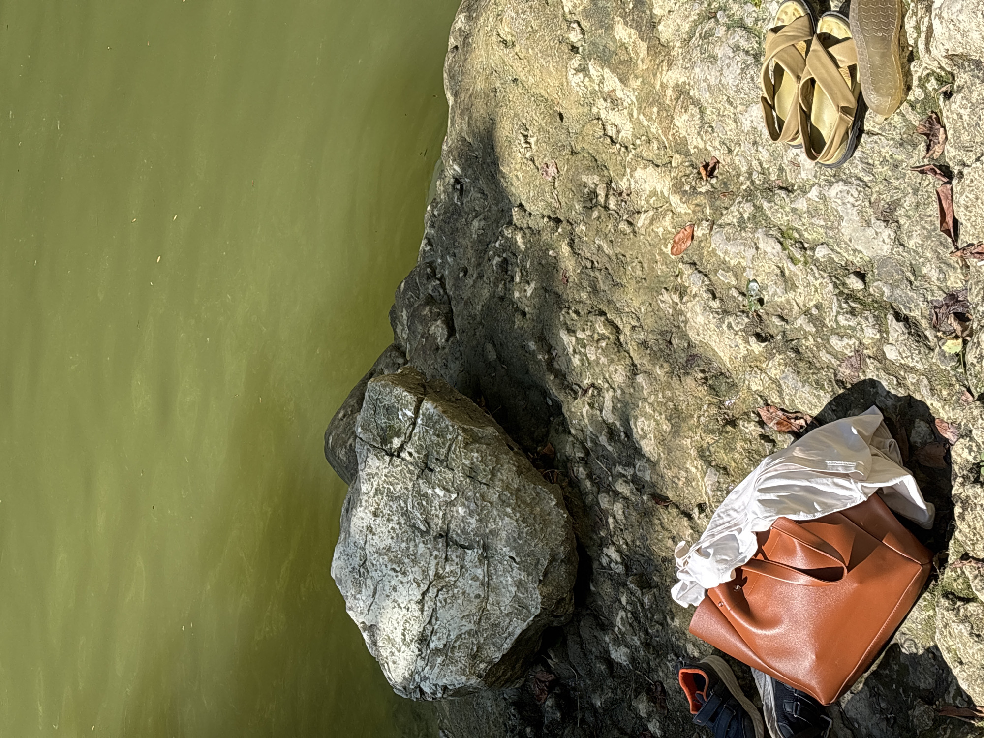 This is a medium shot, eye-level, outdoor, natural light, close-up photograph taken on a rocky shoreline, presumably in the city of Khostinskiy, Russia. The foreground shows a collection of personal belongings laid out on the rocky ground. There is a large, light-colored, rough-textured rock on the left side of the image, adjacent to the water. Next to the large rock, further into the foreground, is a brown leather tote bag, with a white garment peeking out from its opening. To the left of the tote bag are a pair of dark-colored athletic shoes with orange accents. To the right of the tote bag and further into the foreground, there are a pair of tan sandals, and a tan sole of another shoe. The ground is composed of uneven, textured rocks, with sparse patches of dry leaves and some small green foliage. The background consists of a body of murky, greenish water, with subtle ripples on its surface. The water appears calm and reflects the ambient light. The overall scene suggests a moment of pause or rest by the water, with items laid out as if someone has temporarily disembarked or is preparing to do so. There are no people visible in the image.