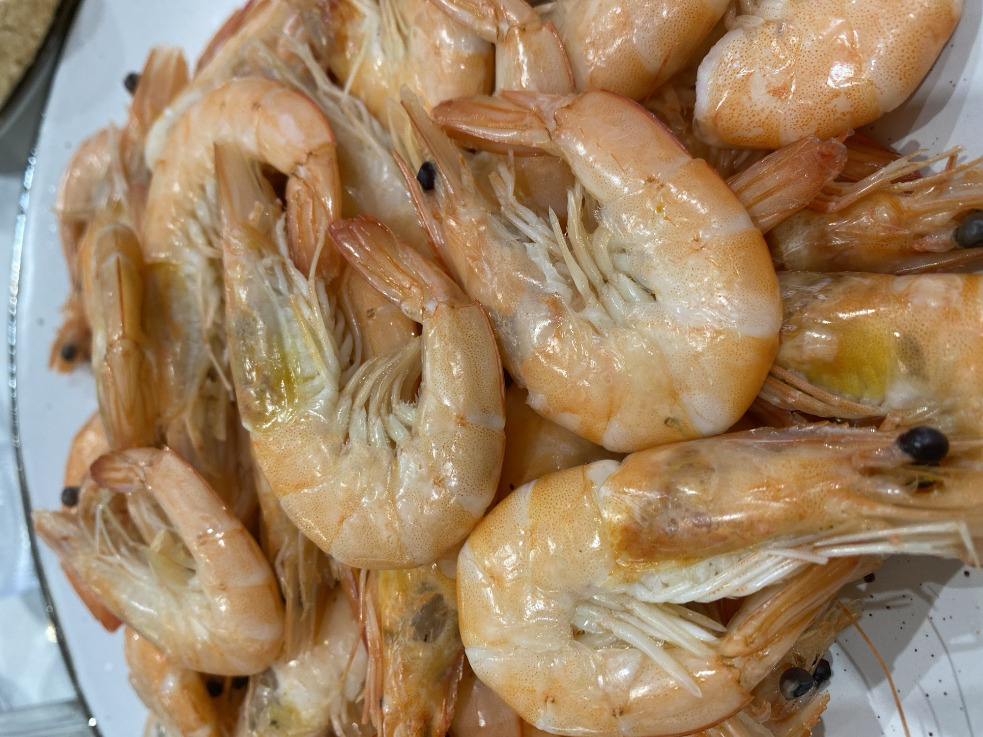 The image shows a close-up of a pile of cooked shrimp on a white plate. The shrimp are pinkish-orange in color and appear to be peeled. Some of the shrimp have their heads and tails still attached. There are a lot of shrimp, stacked unevenly. No location details are available.
