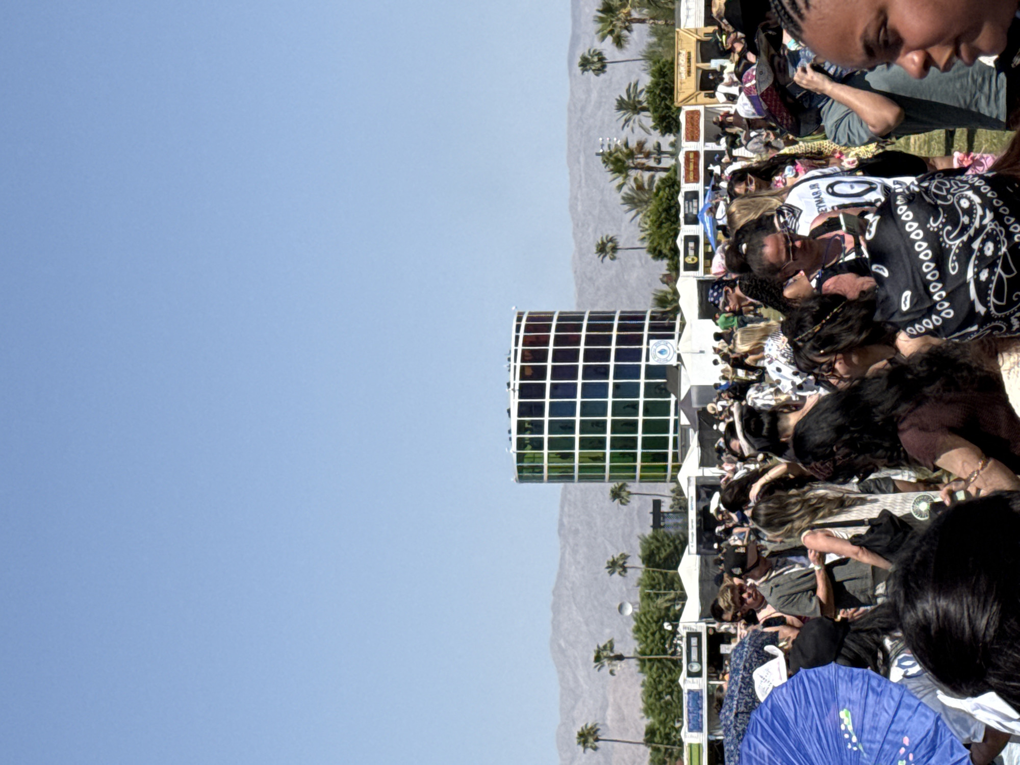 A wide, ground-level shot captures a crowd of people at an outdoor event. In the backdrop, mountains, palm trees, and a clear blue sky are visible. A large, cylindrical structure with a grid pattern and multicolored squares is near the left of the frame, adjacent to the crowd. The event appears to be taking place in the city of Indio, United States of America. The people in the crowd are diverse and engaged in various activities. Some individuals are in the foreground, while others are blurred in the background. Tents and other structures are present amongst the crowd.