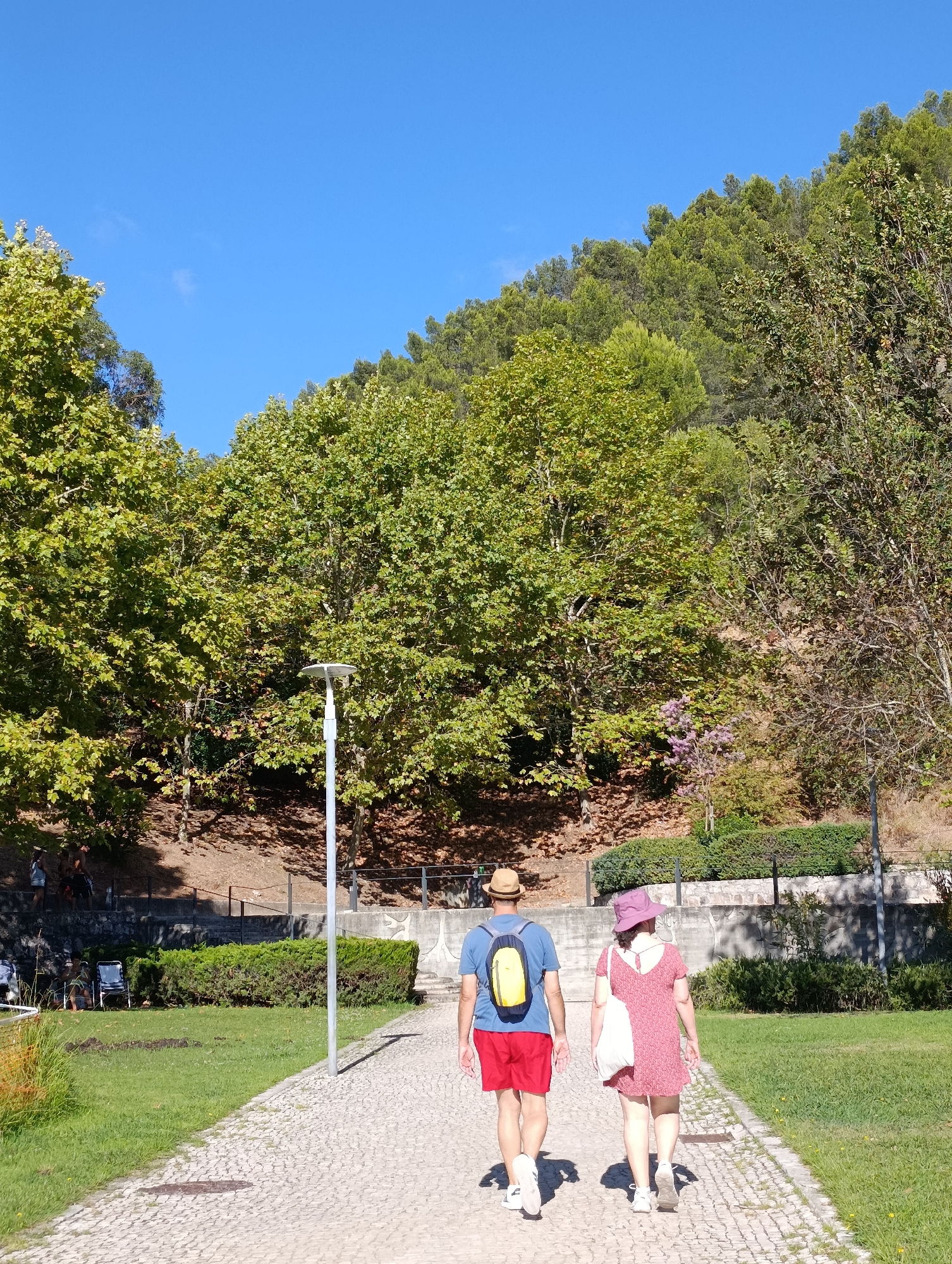 Two individuals, a man and a woman, walk away from the camera on a cobblestone path in a park. The man is wearing a blue t-shirt, red shorts, a straw hat, and a yellow backpack. The woman is wearing a pink dress, a pink hat, and carrying a white tote bag. They are walking towards a lush green hill covered in trees. The sky is clear and blue. In the background, there is a light post and some well-maintained bushes. The park appears to be part of an urban green space, possibly the Parque da Vitalidade in Setúbal, Portugal, given the terrain and vegetation. The overall scene depicts a pleasant day for an outdoor stroll.