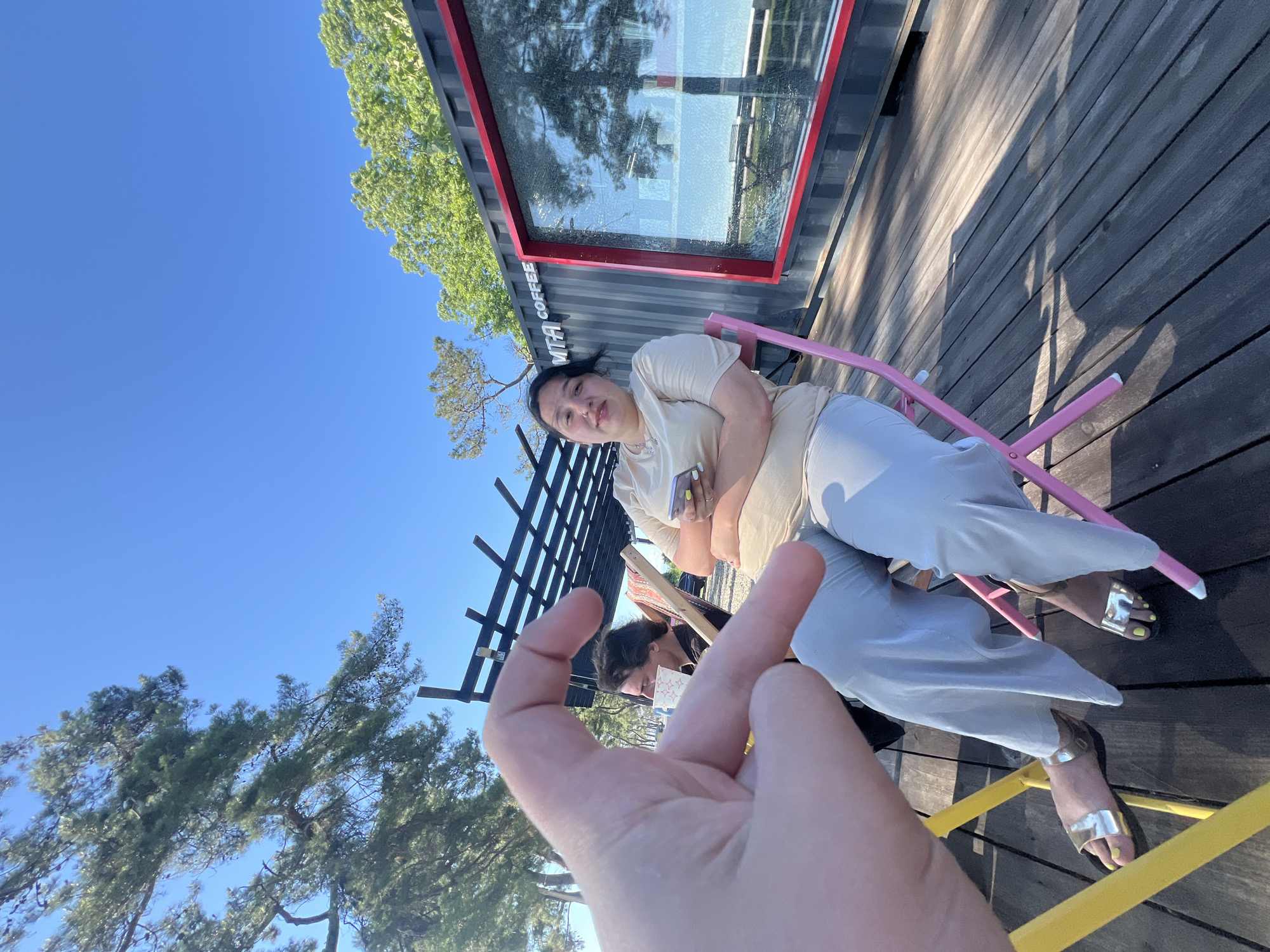 In Ochamchire, Abkhazia, a person's hand is held up in the foreground, forming a heart shape with their fingers. Behind the hand, a woman sits in a pink chair, holding a smartphone. She is wearing a light-colored t-shirt and light-colored pants. To her left, another person sits in a chair, facing away from the camera, holding a white cup. The scene takes place on a wooden deck next to a red-trimmed shipping container with the text "MTA COFFEE" visible. Trees and a clear blue sky are in the background.