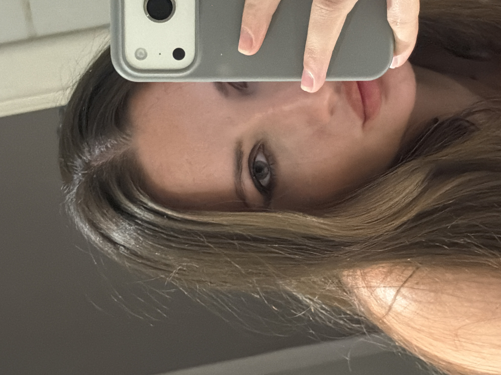 A close-up selfie shows the face of a young woman from the chest up. She is looking directly at the camera, her hair is long and brown with lighter highlights. She is wearing makeup, including eyeliner and lipstick. Her left hand is holding a grey phone in front of her face, obscuring the right side of her face. The background appears to be a bathroom wall, indicated by a slightly reflective surface and the presence of what could be a towel rack or light fixture on the right side. The lighting suggests an indoor setting, possibly illuminated by artificial light. The image does not contain any information indicating it was taken in Sharm El Sheikh, Egypt.