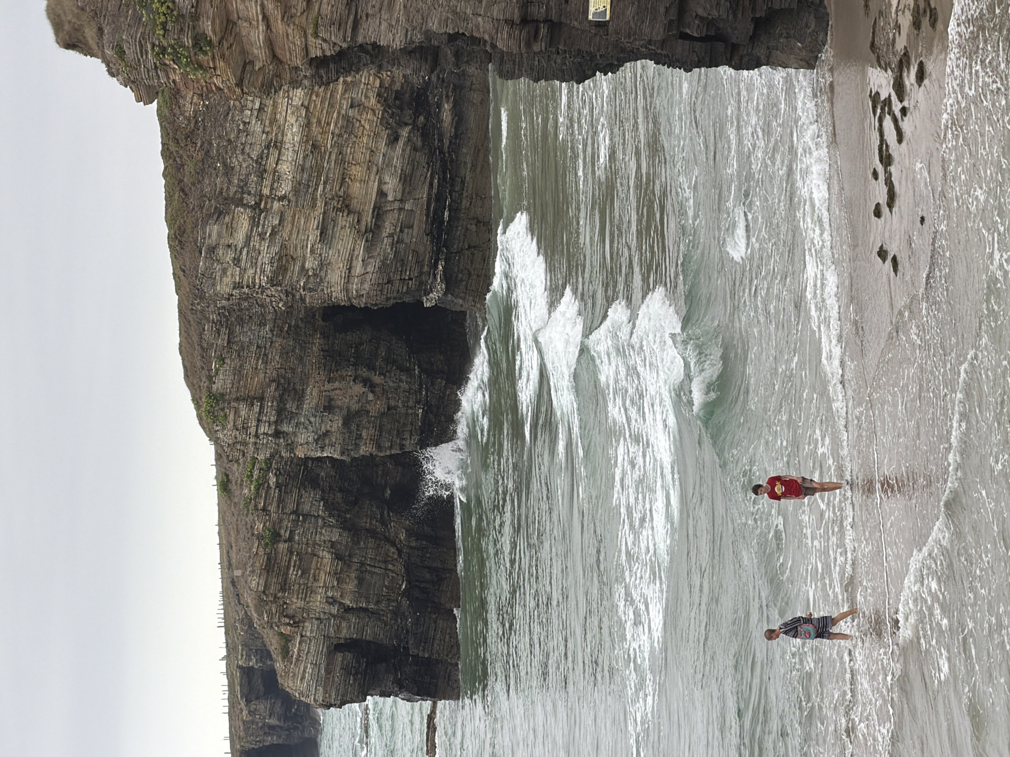 Two individuals are wading in shallow ocean water at the base of massive, layered rock formations. The person on the left, appearing younger, wears a striped dress and carries a bag over their shoulder. The person on the right, also appearing younger, is wearing a red t-shirt and shorts. Gentle waves are breaking around their legs. The rock formations dominate the background, with their stratified surfaces appearing weathered and textured, some showing dark openings or crevices. Vegetation is visible growing on the cliff tops, and the sky is overcast. The scene is consistent with the Praia das Catedrais (Cathedral Beach) in Ribadeo, Spain, known for its striking rock arches and natural sculptures carved by the sea.