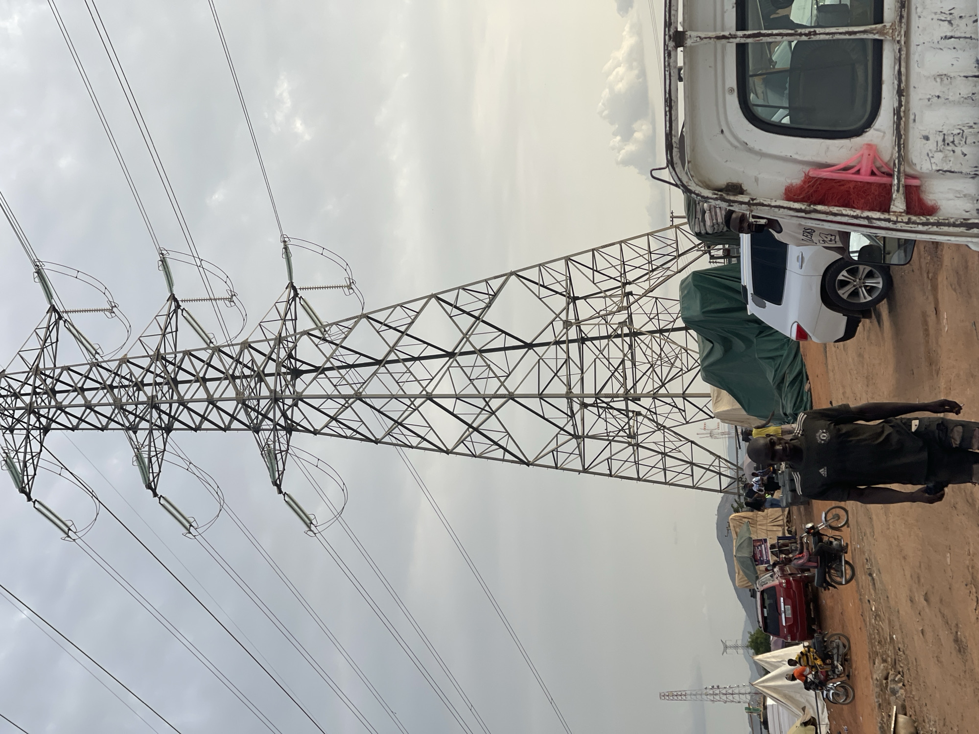 The image shows a bustling scene in Abuja, Nigeria, near a high-tension power line tower. The sky is overcast. The foreground features a tall, lattice-structure power line tower with multiple insulators and high-voltage lines extending towards the sky.
To the right of the tower, there is a white vehicle parked next to a larger, older vehicle (possibly a truck) with a pink broom attached. A man in a black shirt is walking in the lower right corner.
The background includes other power line towers further down the road, tents, parked cars, and motorcycles. People are visible on some of the motorcycles and in the background, suggesting a crowded or market-like environment. The ground appears to be dirt.