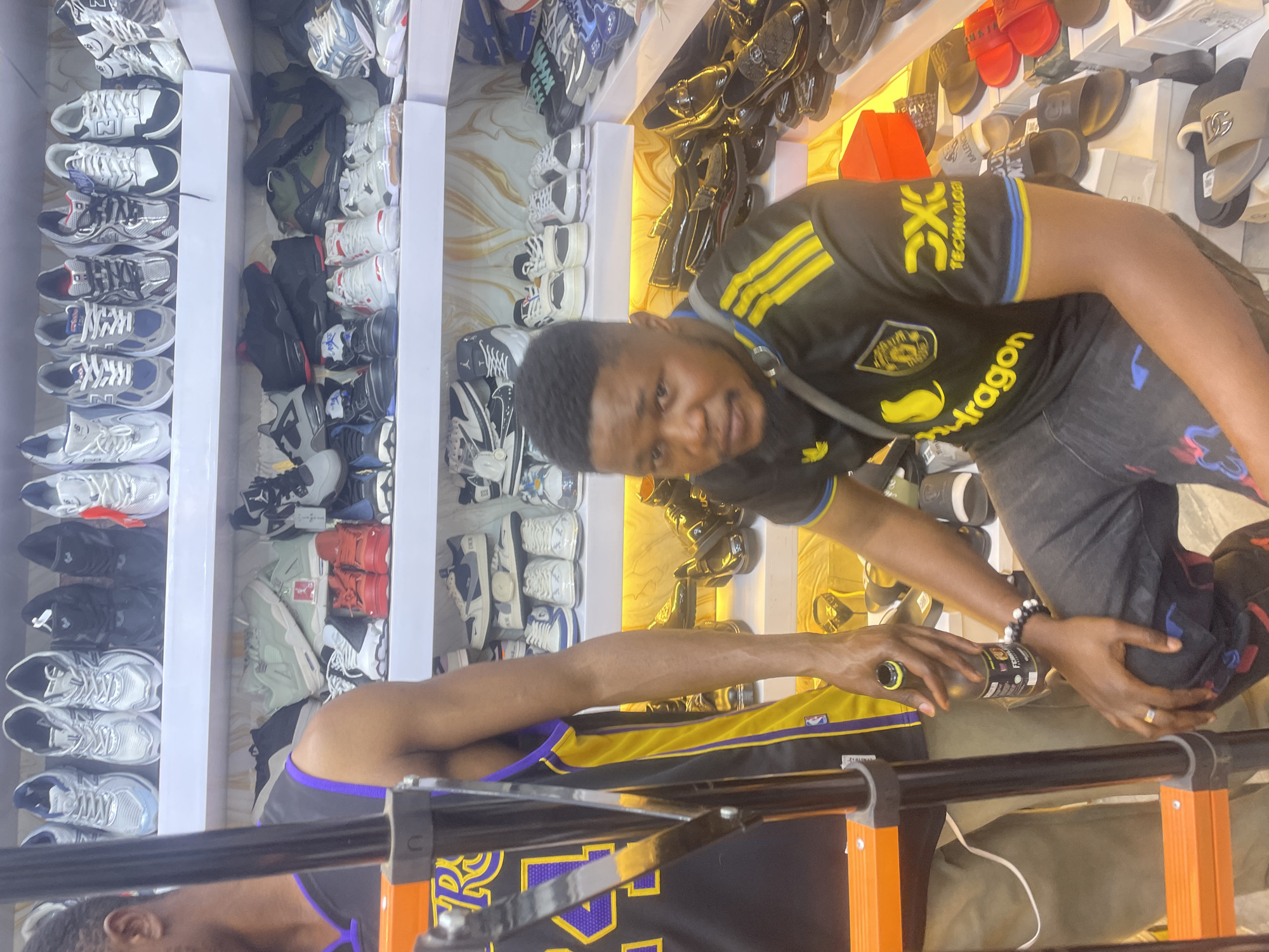 The image shows the interior of a store with shoe displays in Jos, Nigeria. There are rows of shoes and sandals lining the walls. A man wearing a black and yellow jersey is standing in the foreground, partially obscured by what appears to be the top of a ladder with orange supports. He is looking at the camera while holding a bottle. The second person is wearing a yellow and purple Lakers jersey and holding the same bottle as the first man.