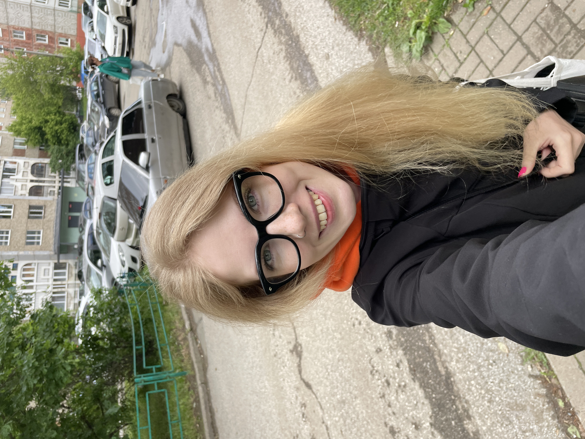 An outdoor selfie captures a smiling young woman with long, straight blonde hair and light-colored eyes, wearing distinctive black-rimmed glasses and a small nose ring. She is dressed in an orange hoodie under a black jacket. Her left hand, adorned with bright pink nail polish, is visible near her hair. The image is rotated 90 degrees clockwise.
The background shows an urban residential setting, consistent with Izhevsk, Russia. Several parked cars, including silver, dark, and white models, line what appears to be a street or parking area. Behind them, multi-story apartment buildings, typical of post-Soviet architecture, feature numerous windows and some weathering on their facades. Lush green trees and patches of grass add natural elements to the scene, along with a low, green metal fence. The ground is paved, showing cracks and what might be damp patches, suggesting recent rain or an overcast day.
The woman is taking a self-portrait, looking directly at the camera with a cheerful expression. The lighting indicates it is daytime, likely without direct sunlight. No discernible text is visible within the image.