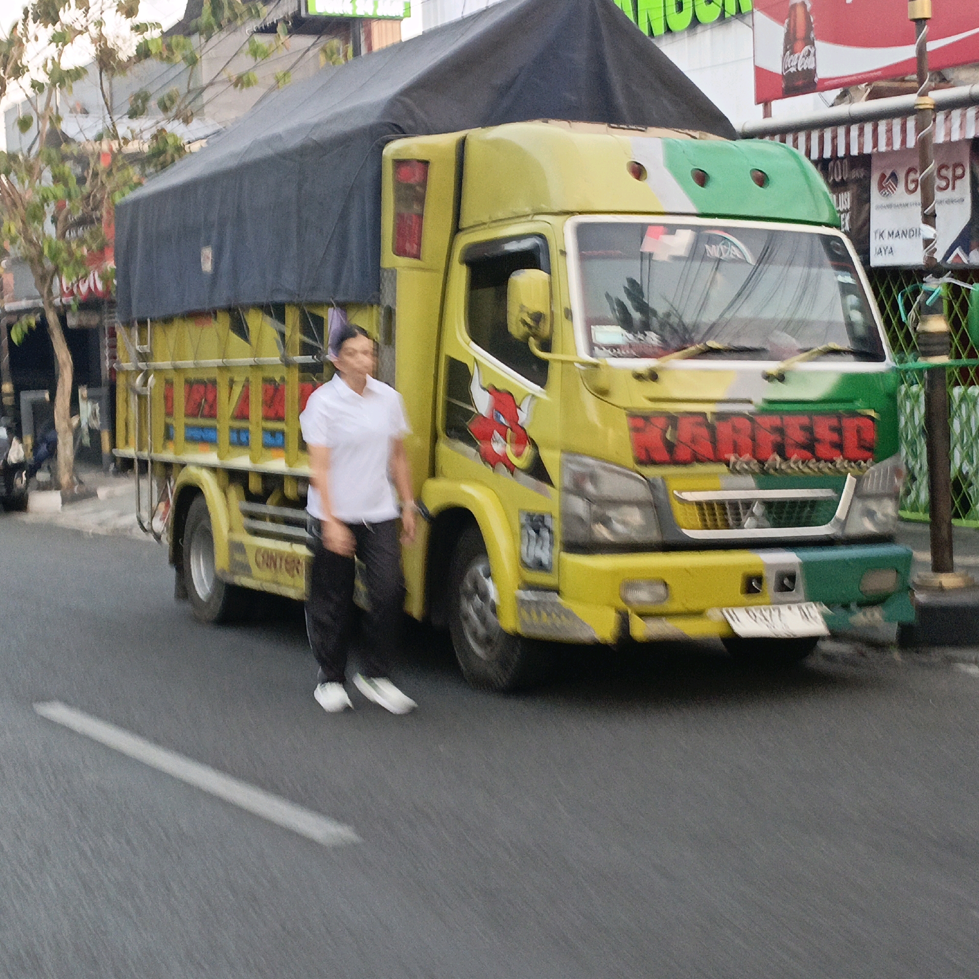 A yellow truck with a dark tarp cover is parked on the side of a street in Salatiga, Indonesia. A person in a white polo shirt and dark pants walks in front of the truck. The truck has graphics on the side and text that appears to say "KARFFED" in red lettering. The environment shows a street with buildings and trees in the background. The image is slightly blurred, indicating motion blur.