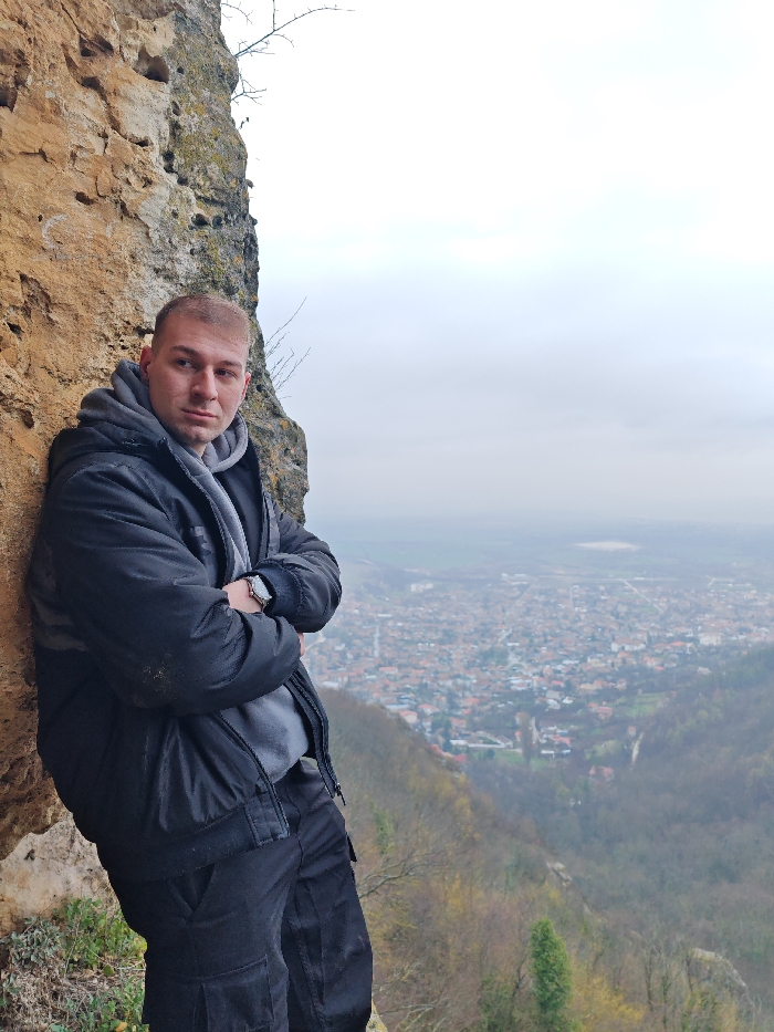 An adult male subject is positioned on the left side of the frame, leaning his left shoulder and back against a textured, light brown rock face. He has short, light-colored hair and a neutral expression, looking directly forward. He is wearing a dark, possibly black, puffer jacket over a grey hooded sweatshirt and dark cargo pants. His arms are crossed at chest level, and a silver-toned wristwatch is visible on his left wrist.
To the right of the subject, a wide panoramic view unfolds under an overcast sky. This view encompasses the city of Shumen, Bulgaria, spread across a valley and low-lying hills. The urban area consists of numerous buildings, primarily residential, extending into the distance. The valley immediately below the viewpoint is heavily forested with deciduous trees, showing sparse foliage consistent with late autumn or winter conditions. The scene suggests a vantage point from a natural rock formation on the Shumen Plateau, offering an elevated perspective of the city below.