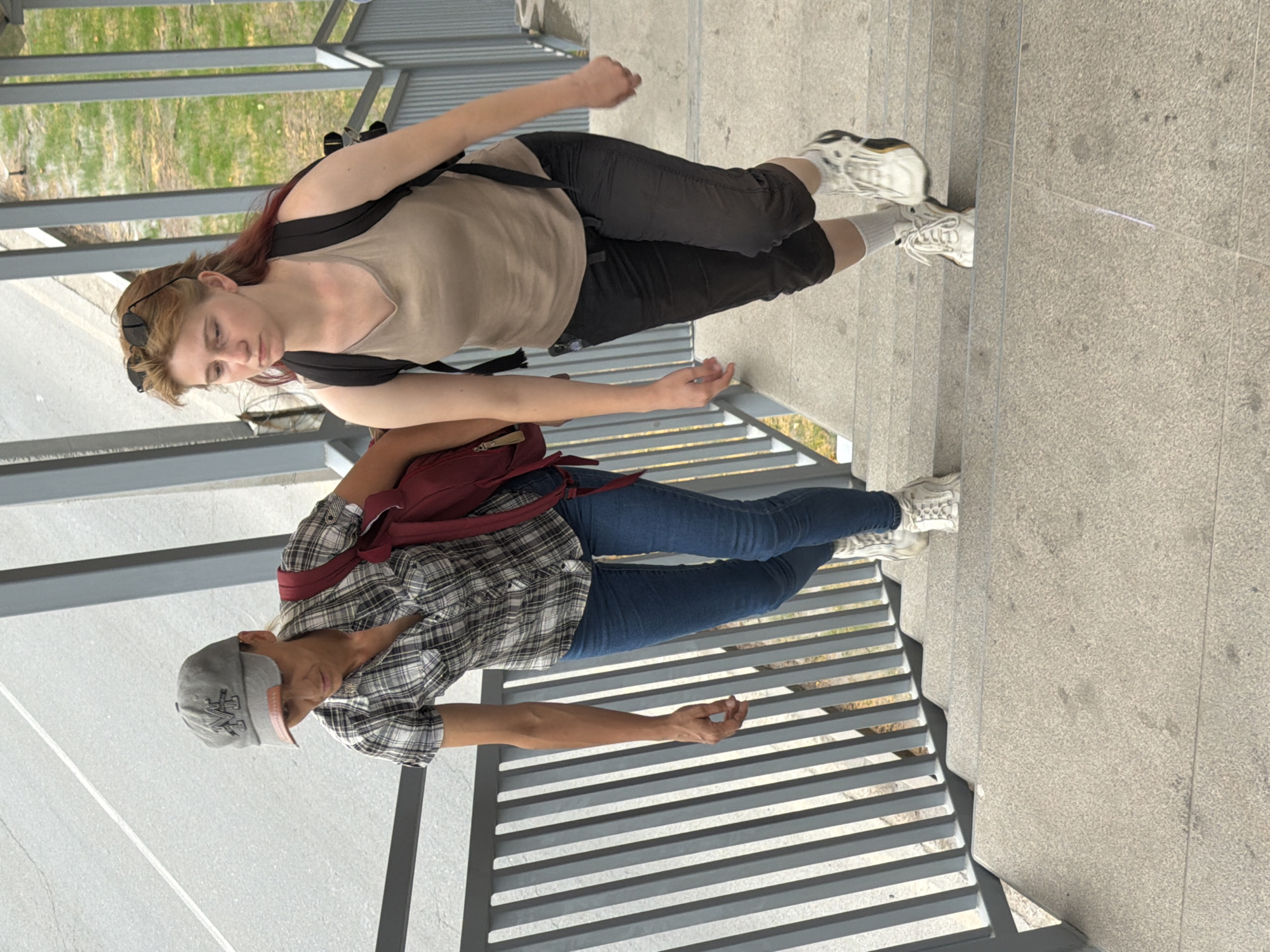 Two individuals are ascending a concrete staircase with metal railings. The person on the left, wearing a grey baseball cap, a plaid short-sleeved shirt, and blue jeans, carries a red backpack. The person on the right, with sunglasses perched on their head and hair with red highlights, wears a beige tank top and black capri pants and carries a black backpack. Both individuals are wearing athletic shoes and white socks. The background shows a paved area and greenery.