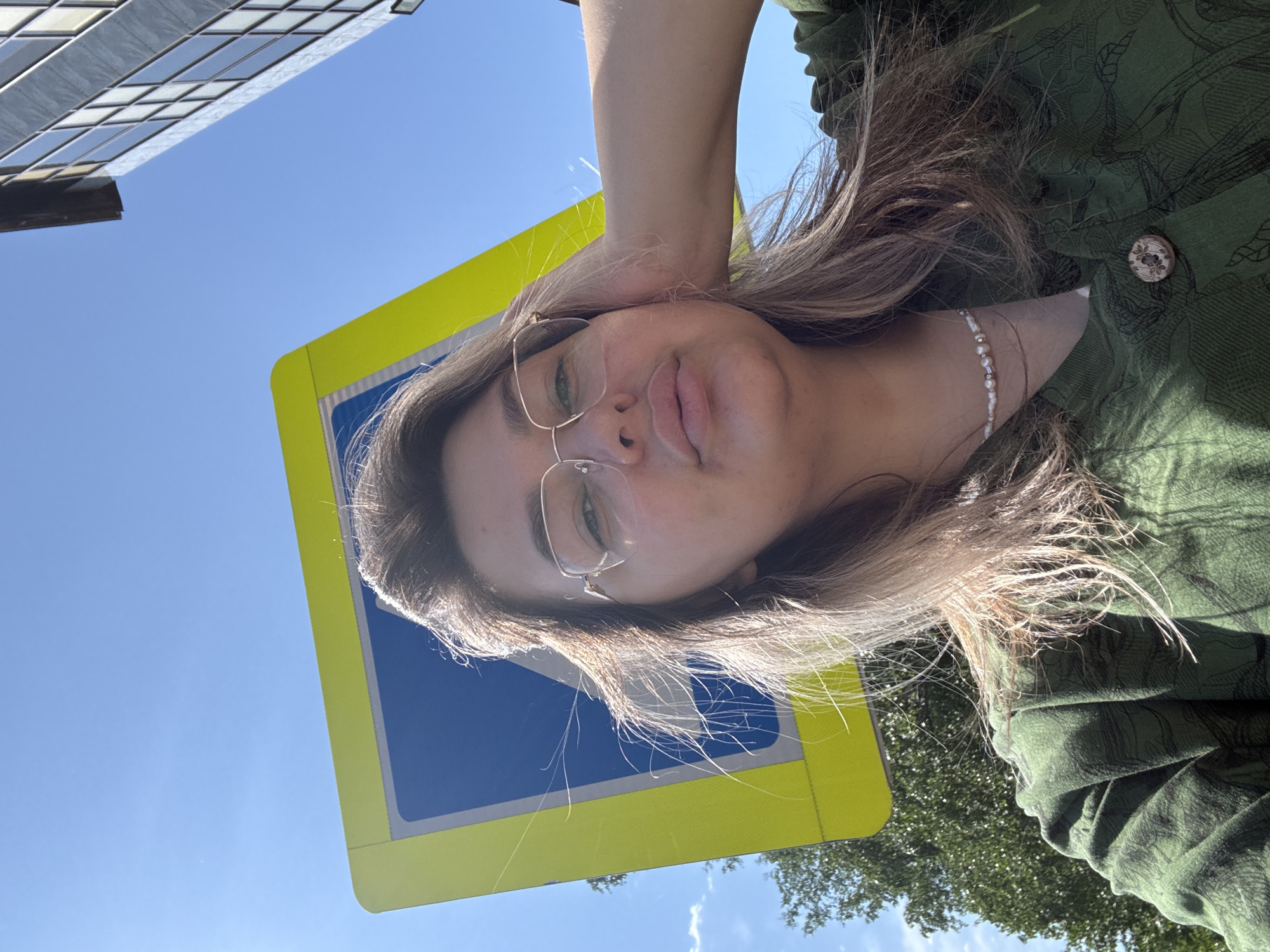 A woman with blonde hair and glasses is taking a selfie outdoors. She is wearing a green jacket with a pattern and a beaded necklace. Behind her is a blue and yellow pedestrian crossing sign and a modern building. The sky is clear and blue. The image appears to have been taken in Birobidzhan, Russia, based on external context.