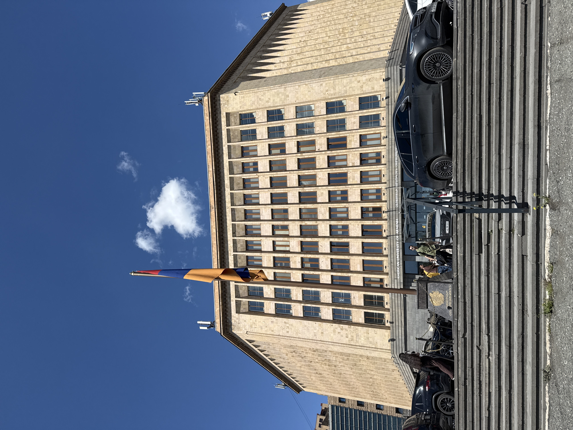 The image shows the American University of Armenia (AUA) in Yerevan, Armenia. The building is a large, multi-story structure with numerous windows and a pale, textured facade. The Armenian flag flies in front of the building against a clear blue sky with a few clouds. Stairs lead up to the entrance, and several people are on the stairs. Two dark-colored cars are parked nearby. A metal railing runs along the side of the stairs. A plaque is visible on the stairs. The overall setting is urban and appears to be a sunny day.