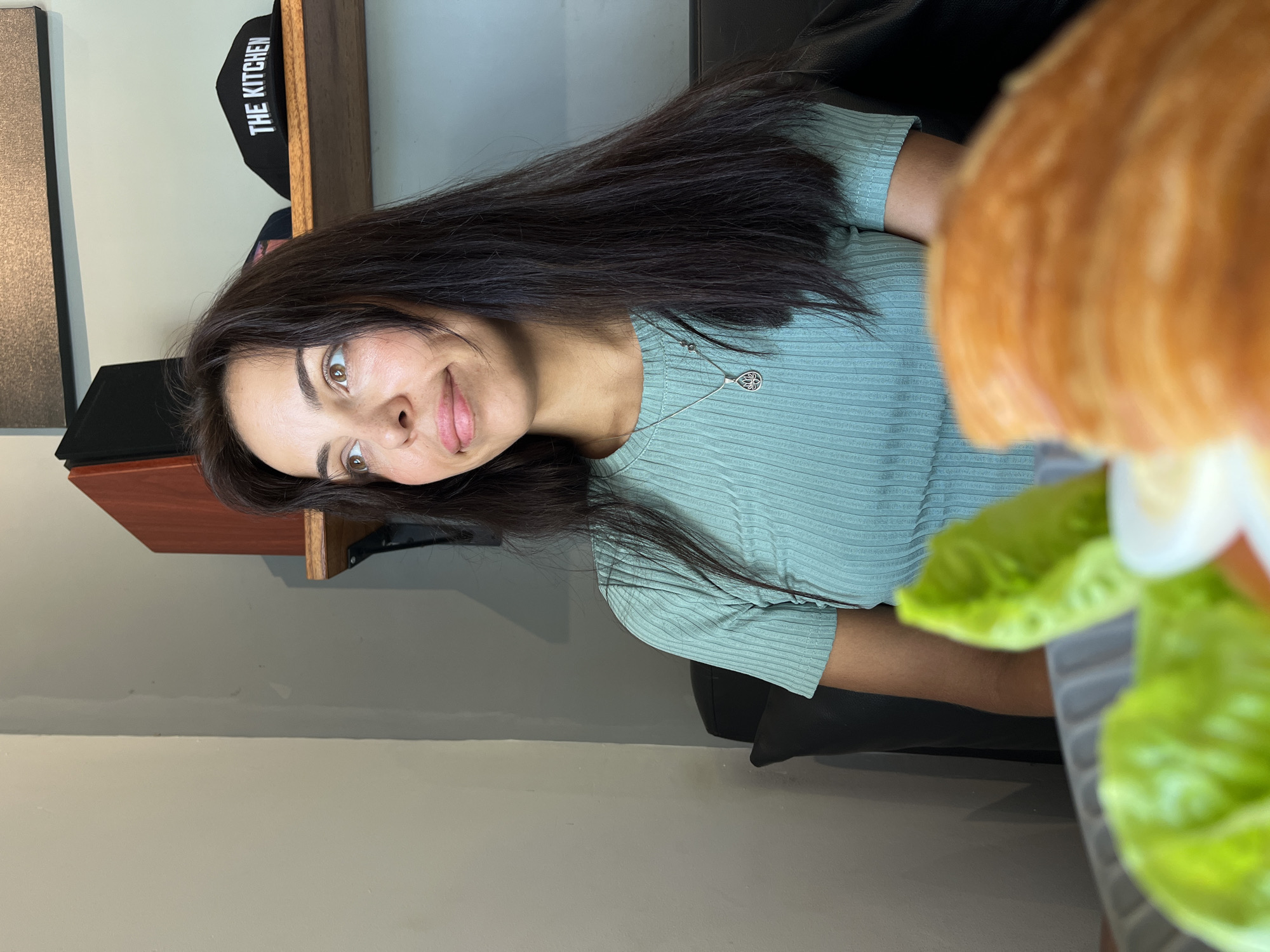 A young woman with long, dark hair is seated indoors, smiling slightly at the viewer. She wears a light green ribbed top and a delicate silver necklace. A plate of food is partially visible in the foreground, containing lettuce, a hard-boiled egg, and possibly bread or pastry. Above her and slightly behind her head, there is a shelf with a black baseball cap labeled "THE KITCHEN". The background shows a plain wall and what looks like a black leather chair. There are no elements in the photograph that would suggest the location is specifically in Kuta Selatan, Indonesia. The description is based solely on visual details.