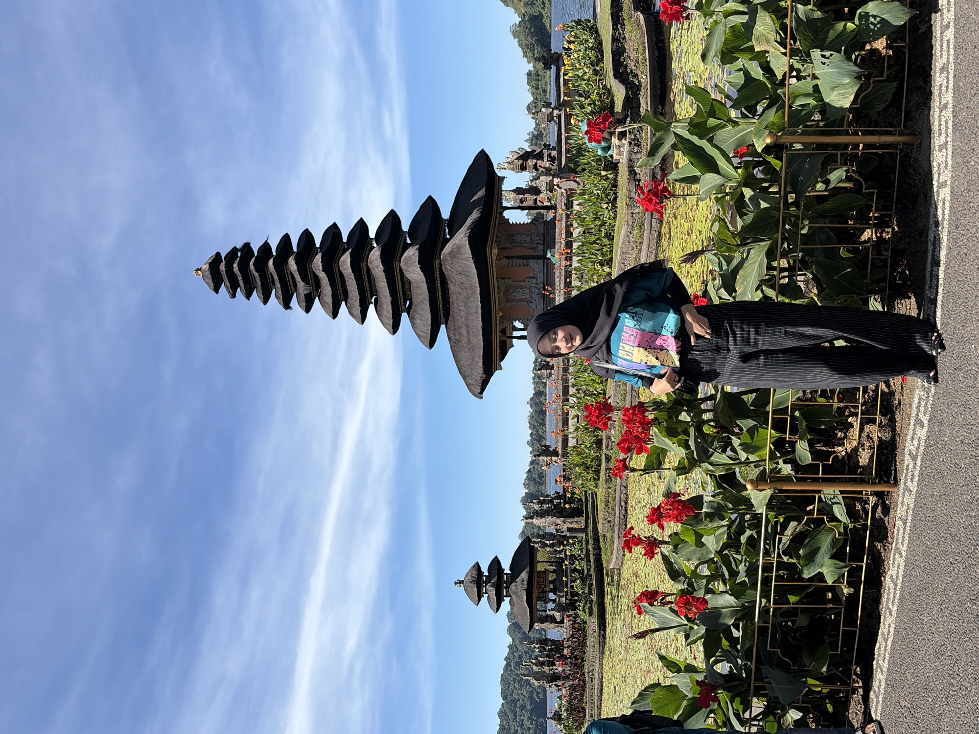 A woman wearing a black hijab and a colorful t-shirt is standing in front of a serene lake with lotus pads and a traditional Balinese temple. The temple, known as Pura Ulun Danu Beratan, is located in the city of Baturiti, Indonesia, and is characterized by its tiered roof structure. The woman is looking away from the camera with a slight smile, and her hands are placed in her pockets. She is wearing black and white striped pants. In the background, the lake is surrounded by lush greenery and hills. The sky is clear and blue with scattered clouds, indicating a sunny day. The scene conveys a sense of tranquility and cultural richness.