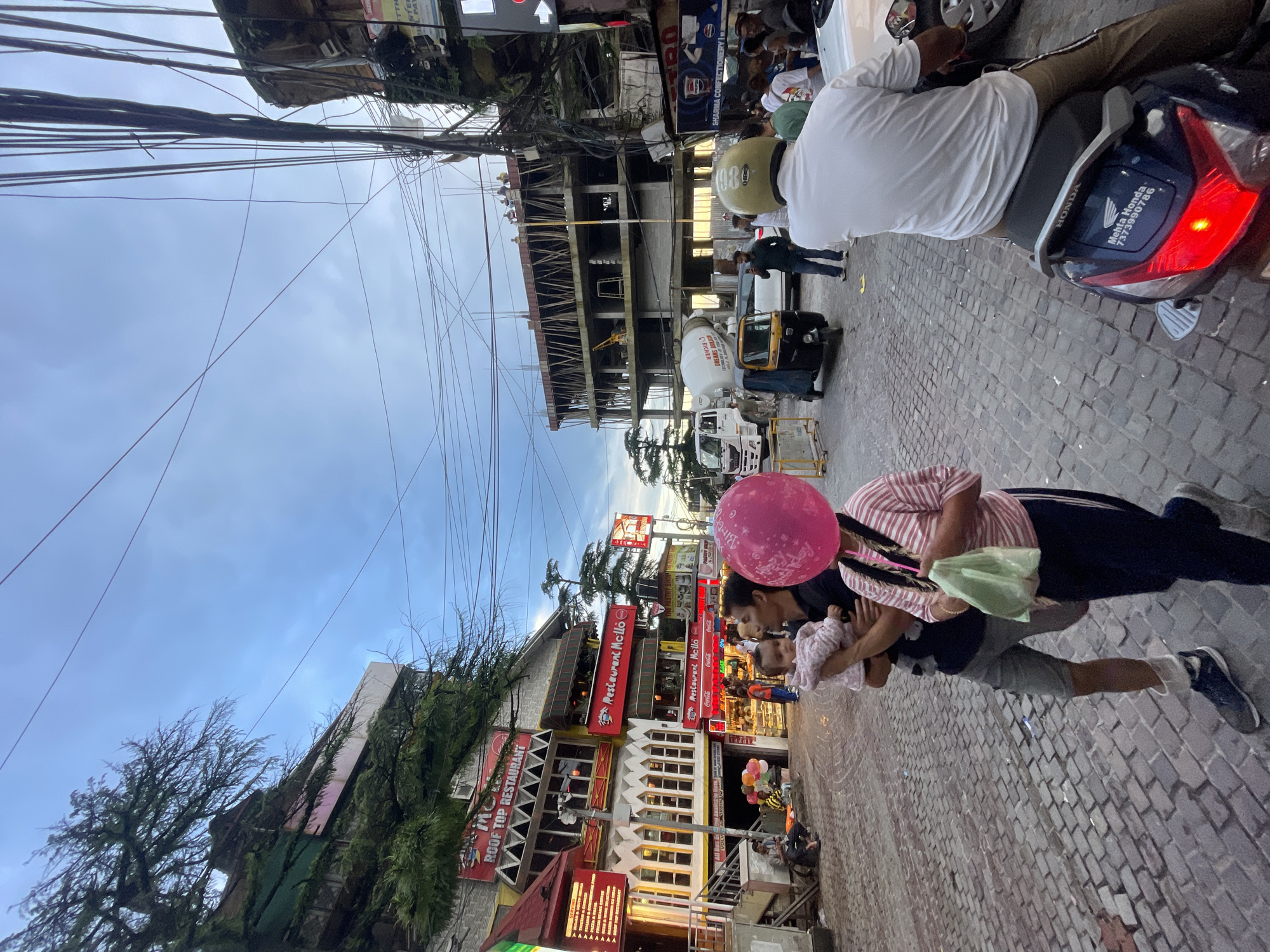 In Bhagsu, India, a street scene unfolds beneath a cloudy sky. The cobblestone street is lined with buildings, some of which feature vibrant signage for restaurants and shops, including "ROOF TOP RESTAURANT" and "Restaurant Mello." A multi-story building under construction is visible in the background, with workers present on the upper levels. A cement mixer truck and an auto-rickshaw are also on the street.
In the foreground, a family is walking. A man carries a baby. A woman walks beside them, holding a large pink balloon that obscures her face. Another person on a motorbike is seen from behind, with the "HONDA" logo visible on the vehicle. The scene is bustling with activity, suggesting a public gathering or a busy market area.