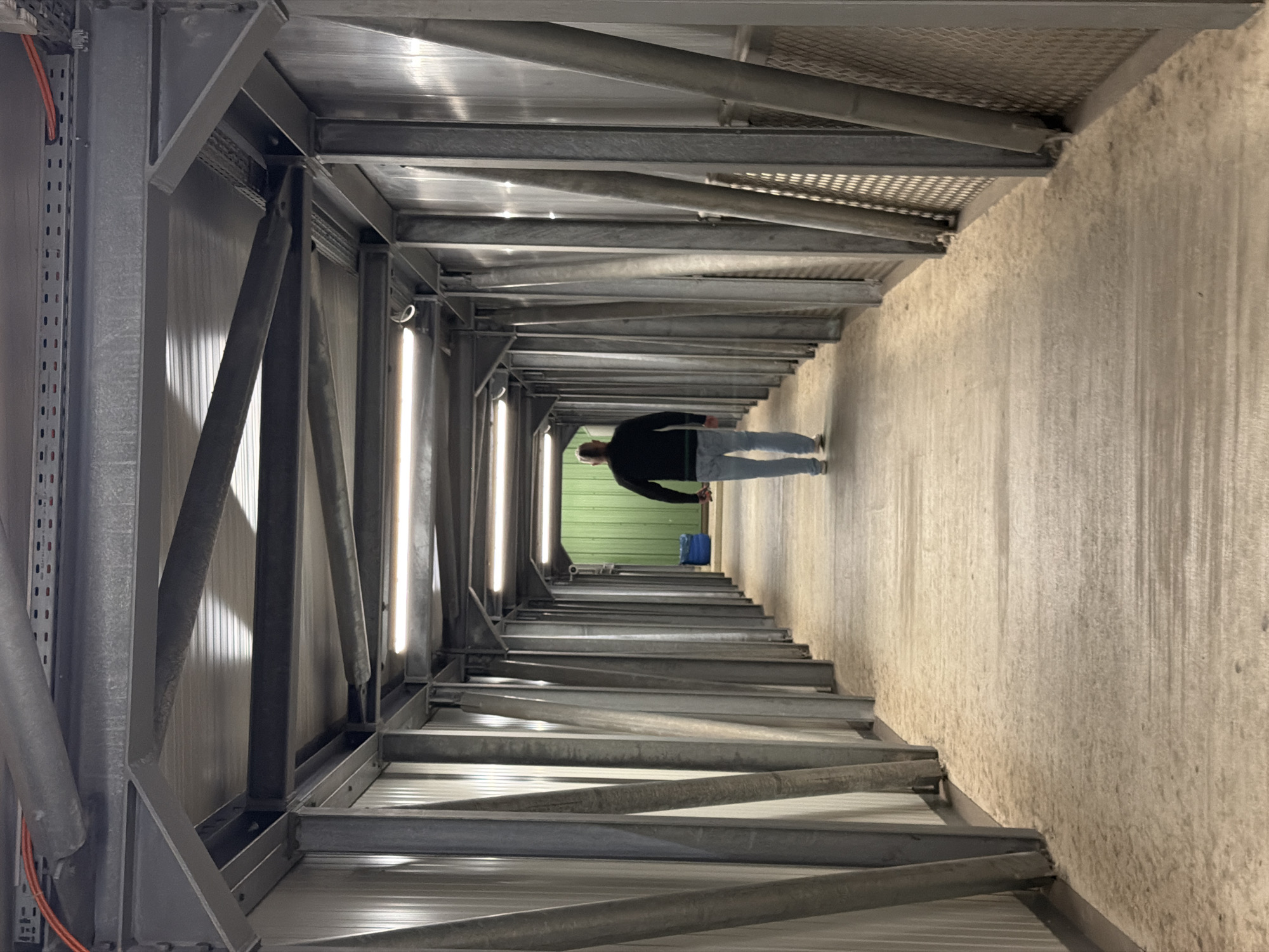 A man walks down a long, industrial-style corridor towards a green wall. The corridor is enclosed by metal structures and lined with vertical metal supports on either side, creating a tunnel effect. The floor appears to be covered in a light-colored granular material, possibly sand or dust. Fluorescent lights are mounted on the ceiling, illuminating the walkway. On the left side of the corridor, near the end, a blue cylindrical object, possibly a bin, is visible. The man is wearing a dark long-sleeved shirt and light-colored jeans, and is walking away from the camera. The overall environment suggests a utilitarian or manufacturing setting. The image is a photograph taken in Leudelange, Luxembourg, likely within an industrial or logistical facility.