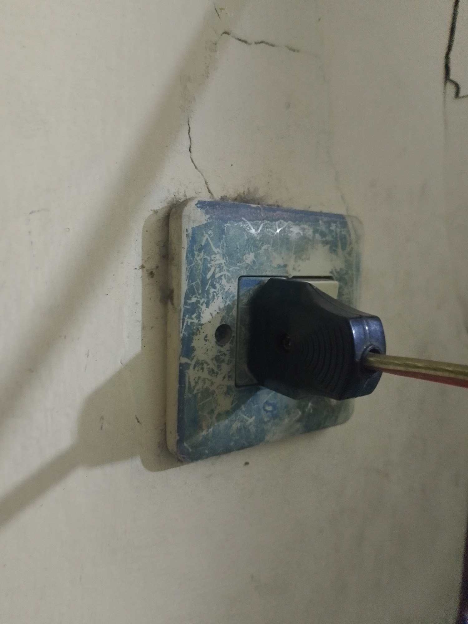 A close-up, eye-level shot shows a black electrical plug inserted into a light blue and white, heavily scratched wall socket. The wall surrounding the socket is a textured off-white, with a noticeable crack running diagonally from the upper left towards the socket. A faint shadow is cast on the wall to the left of the socket, suggesting a light source from the right. A thin red and yellow wire extends from the plug. The scene is indoors, likely in a residential setting in Mukapayung, Indonesia, given the context. There are no people visible in the image.