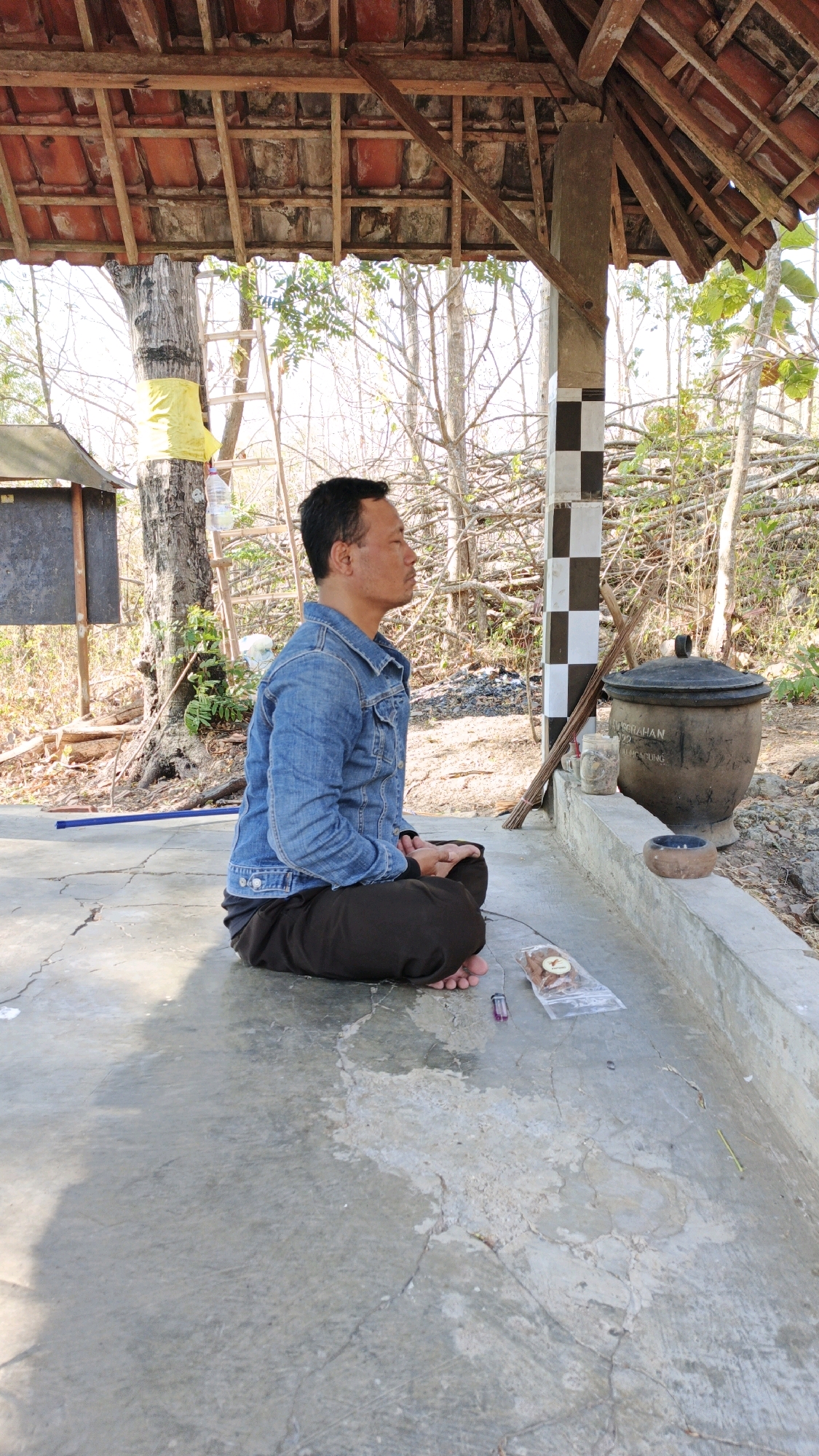 A man sits cross-legged on a concrete floor beneath a rustic, open-air structure with a tiled roof. He wears a denim jacket and dark pants, with his eyes closed in meditation. To his right, a large, dark clay pot rests on a raised concrete ledge, along with some incense sticks and a small bowl. The background shows sparse vegetation and trees, suggesting an outdoor setting in a rural or semi-rural environment. The overall impression is one of quiet contemplation in a simple, outdoor location. The text on the pot may indicate it is located in Junjung, Indonesia, but this cannot be confirmed from the visual information alone.