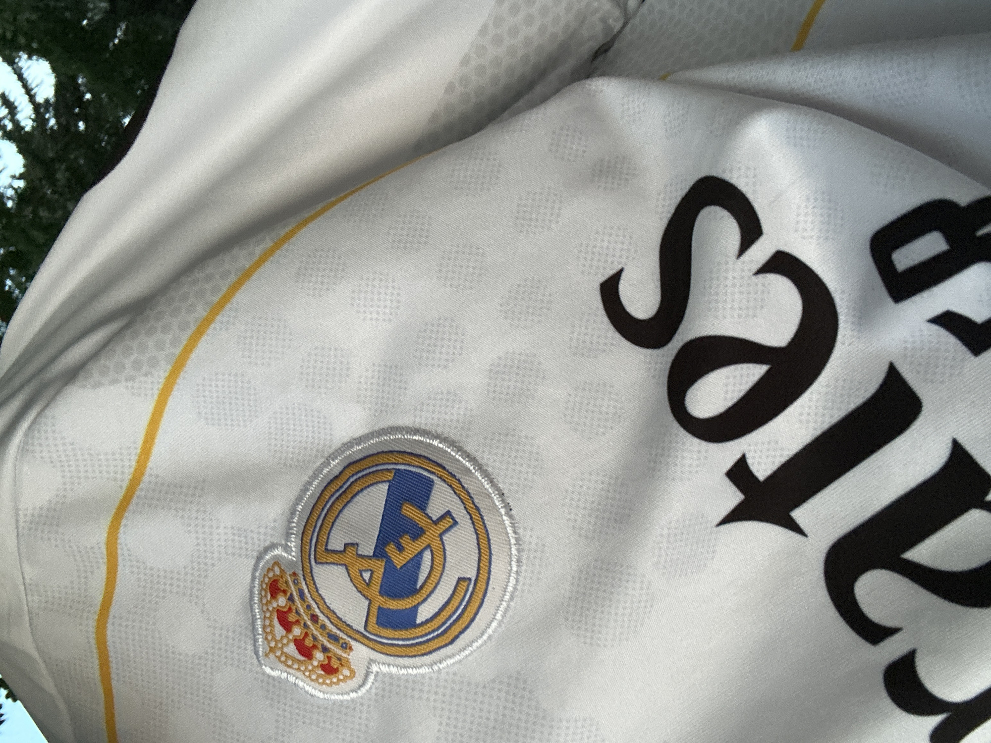 The image shows a close-up of a white Real Madrid jersey with a yellow stripe across the chest and the team's crest. The jersey has a textured pattern of small hexagonal shapes. A portion of the word "Emirates" is visible in large, black, serif font across the lower part of the jersey, indicating it's a sponsor's logo. The background is blurred and appears to be outdoor foliage, suggesting the photo was taken outside. There is no information in the image to suggest a location in Ognianovo, Bulgaria.