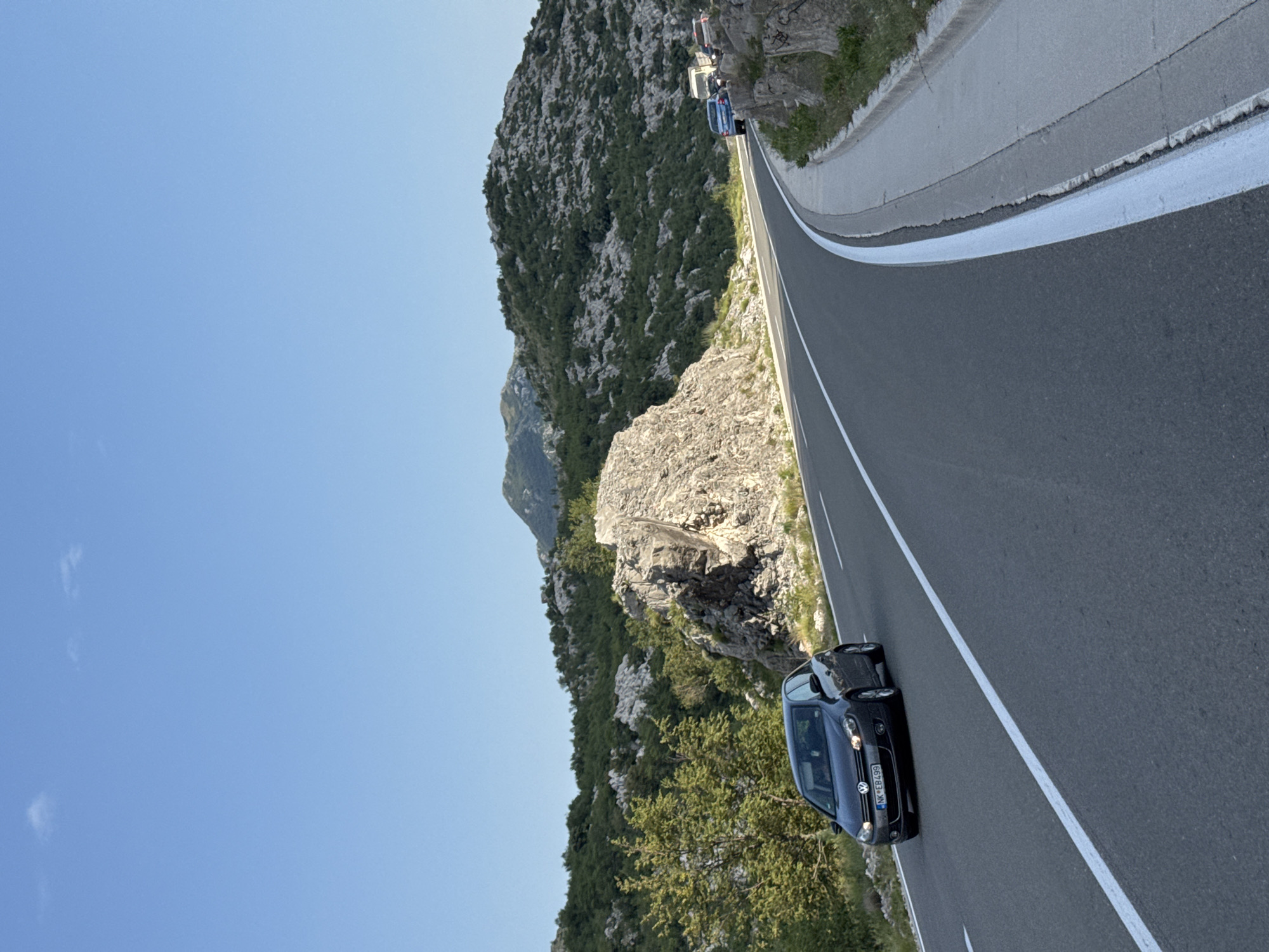 The image depicts a winding road carved into a rocky, mountainous landscape. The road has a grey asphalt surface with a white lane marker. A dark grey Volkswagen Golf is driving on the road in the foreground, with its license plate visible as "NK*EB499". To the right of the road, a concrete sidewalk or barrier runs alongside the edge. In the background, lush green vegetation covers the undulating hills and mountains, with sparse rocky outcrops and boulders interspersed. The sky is a clear, bright blue with a few wispy clouds. The scene appears to be a scenic route, possibly a coastal highway, in the vicinity of Risan, Montenegro, given the characteristic karst topography and Mediterranean vegetation. The overall impression is one of a clear, sunny day on a journey through a natural, mountainous environment.