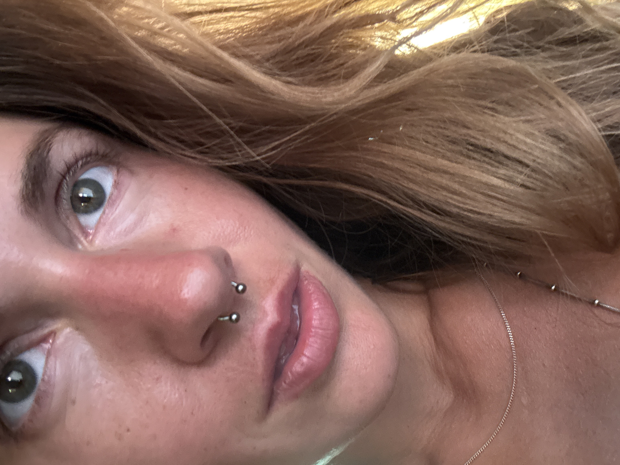 This is a close-up, angled portrait of a young person with blonde hair. The subject has a septum piercing with a horseshoe-shaped barbell, and their eyes are open, looking upwards and slightly to the left. The left eye is visible, showing a green iris. The right side of their face is also visible, featuring their nose and mouth. Their lips are slightly parted and appear to have a pink, glossy finish. A delicate silver chain necklace is visible around their neck and extends towards the right side of the frame. The lighting suggests it might be daytime, possibly with natural light, as there are bright highlights, particularly in the hair and on the skin. The background is filled with the subject's blonde hair, which is slightly wavy. The overall mood conveyed by the subject's expression is one of curiosity or slight surprise. There is no visible text in the image.