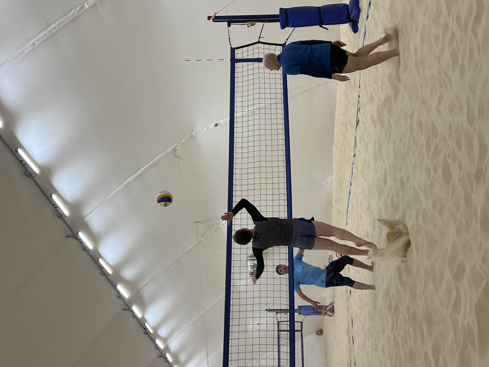 Here's a description of the image:
**Location:** Indoor beach volleyball court, likely Saint Petersburg, Russia.
**Environment:** The court is situated inside an inflatable structure with a white ceiling lit by fluorescent lights. The playing surface is light-colored sand.
**Subjects:** Three adult men are engaged in a beach volleyball game. One man is positioned near the net, reaching for the volleyball with outstretched arms. Another man is slightly behind him, also in a ready stance. A third man is on the opposite side of the net.
**Objects:** A volleyball net with blue supports divides the court. A volleyball is in mid-air above the court.
**Actions:** The men are actively participating in a beach volleyball match, with one player attempting to hit the ball over the net.
**Interactions:** The players are interacting with each other and the volleyball within the context of a competitive volleyball game.
